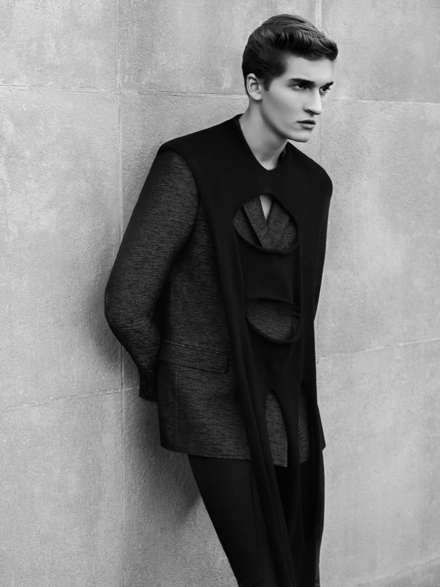 Black-and-white image of a male model leaning on a stone wall in a dark cutout tabard over a tweed jacket, for Bon.