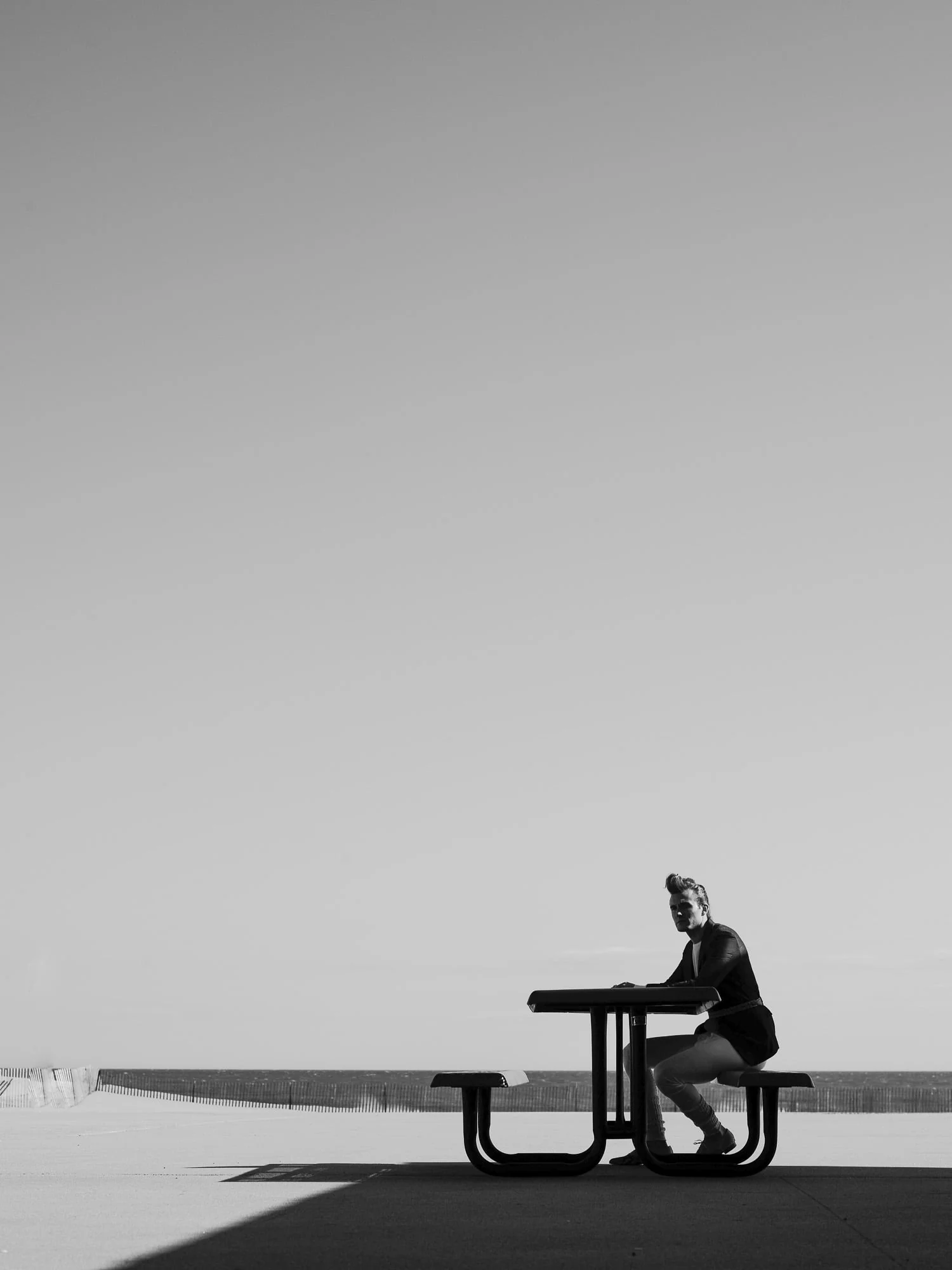 Black and white portrait of Brad Kroenig sitting at a picnic table on a beach boardwalk under a wide empty sky.