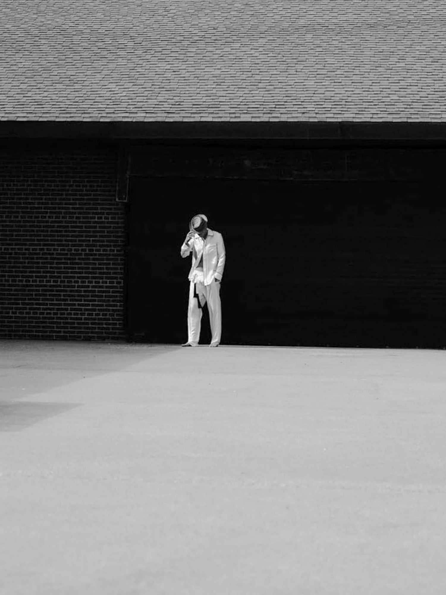 Black-and-white image of Brad Kroenig in a white suit and hat, head bowed, standing before a dark brick wall.