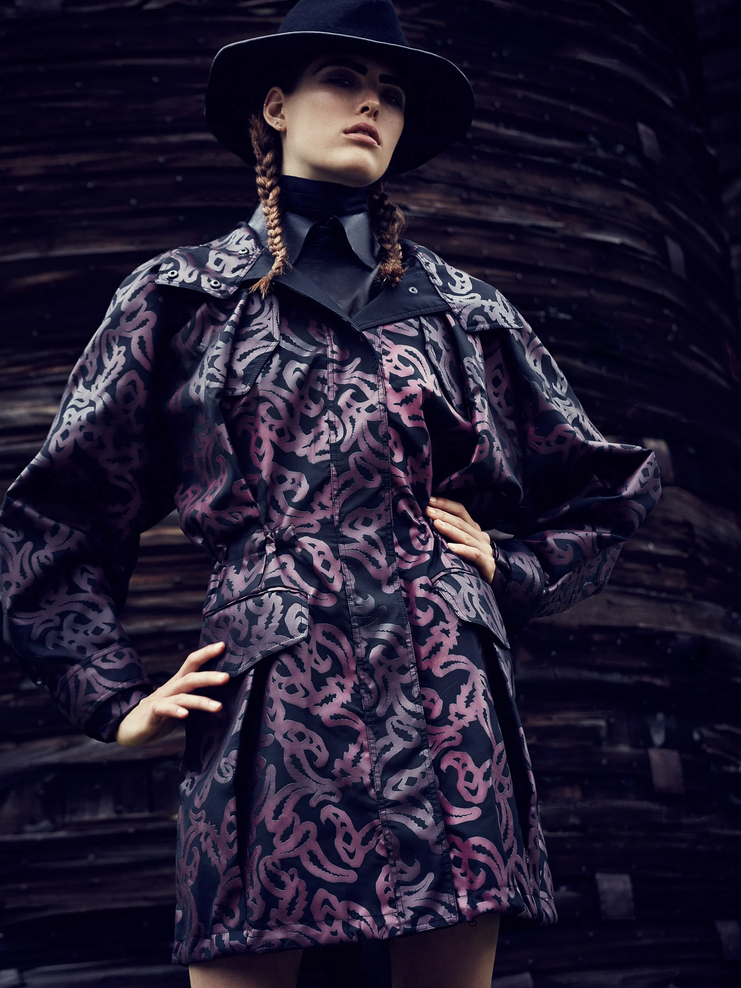 Model with braided hair in a black hat and purple-patterned black coat, hands on hips before a weathered wooden wall.