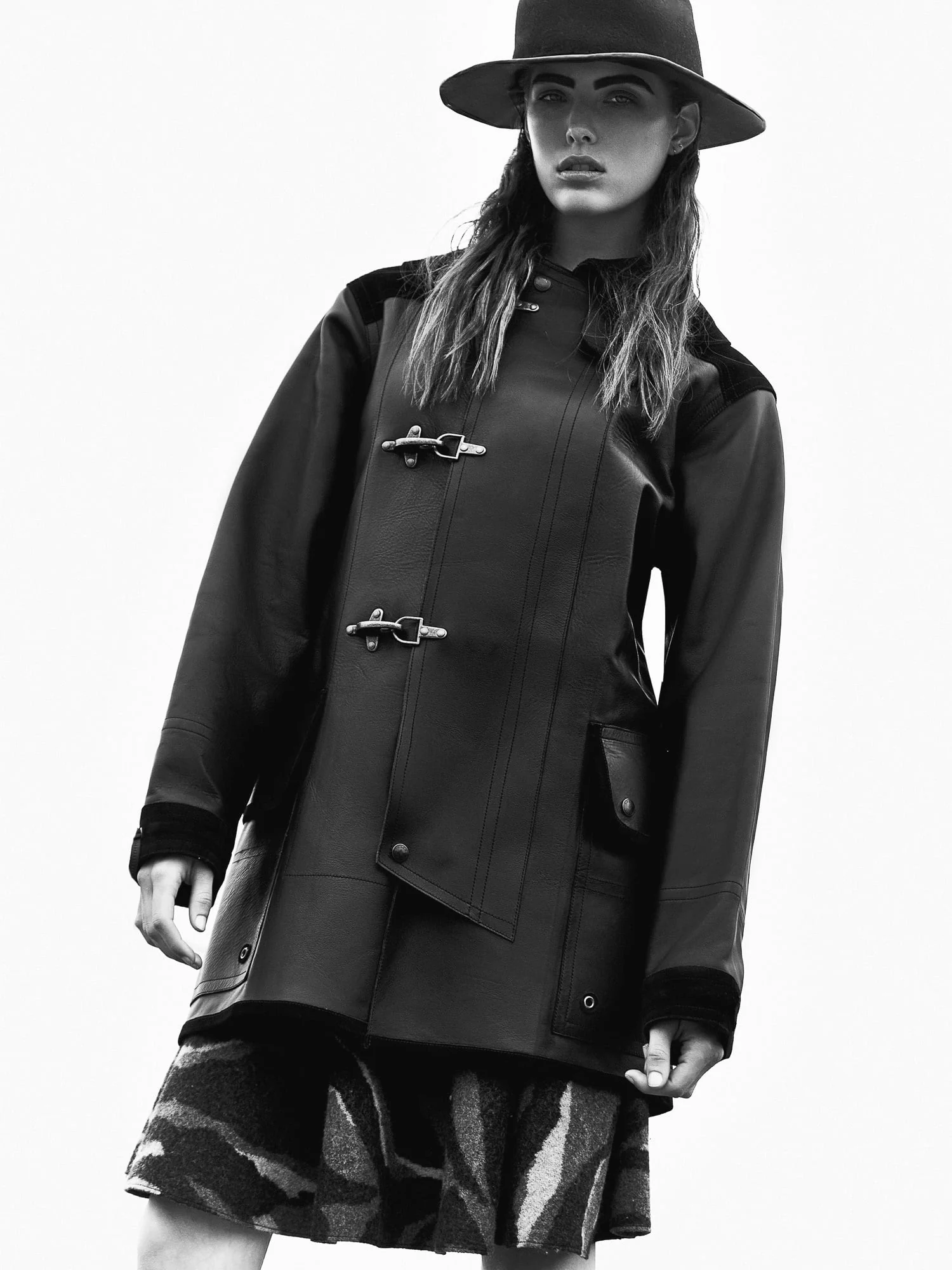 Black and white portrait of Brittany Burke in a leather toggle coat, wide-brim hat and patterned skirt.
