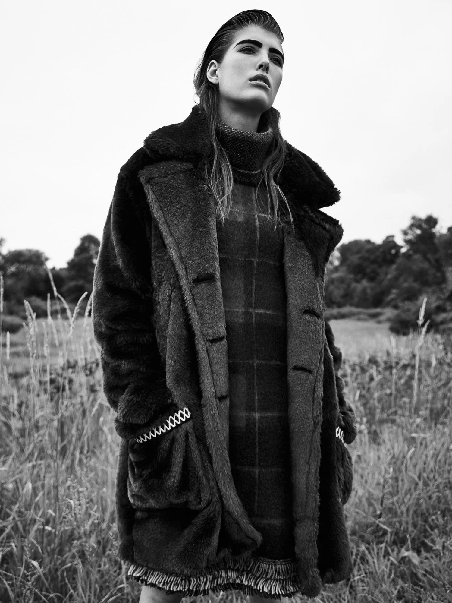 Black and white portrait of model Brittany Burke in a shaggy fur coat over a plaid knit dress, standing in tall grass.