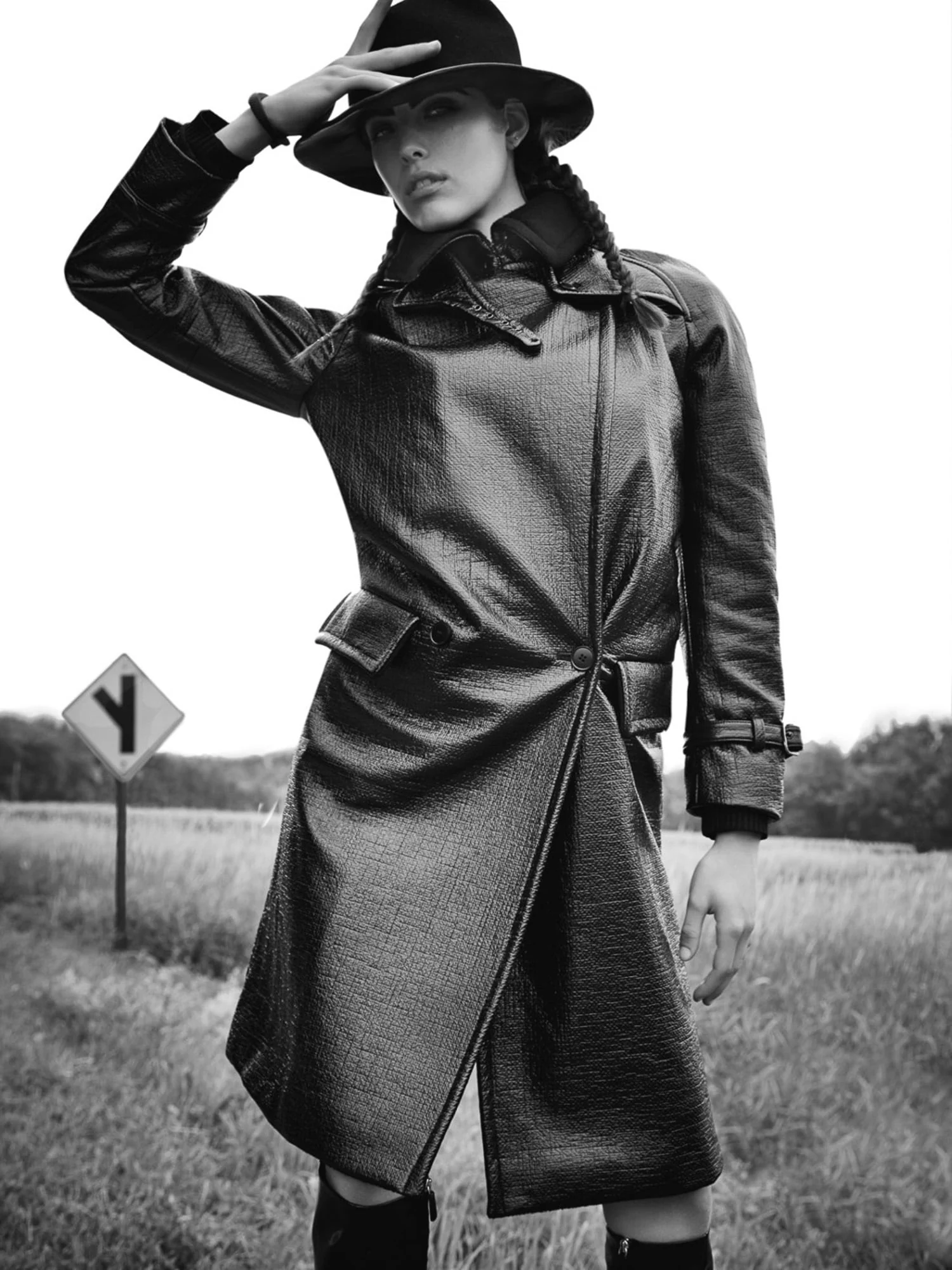 Black-and-white shot of a model in a textured leather coat and wide-brim hat in a grassy field beside a road sign.
