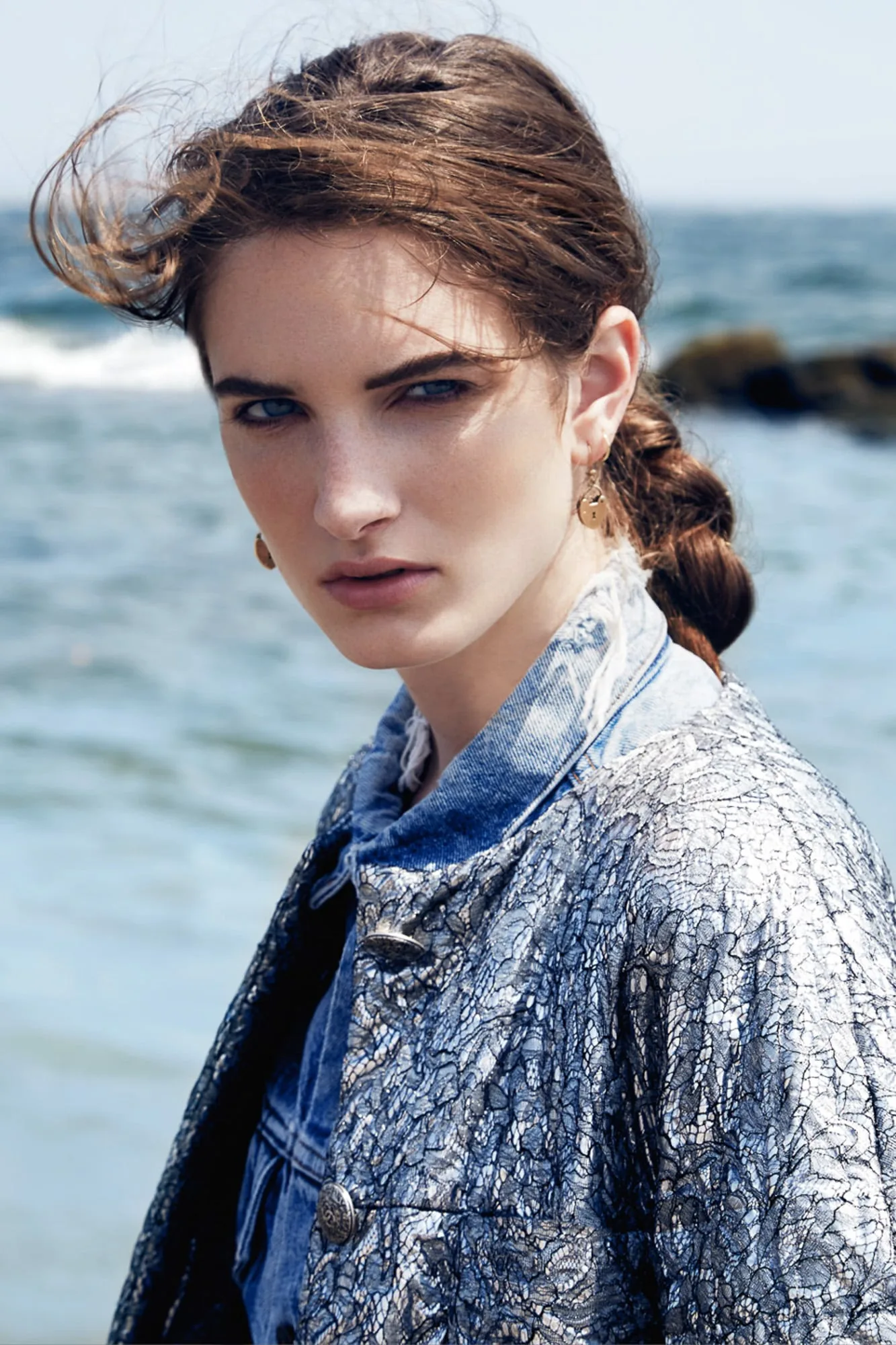 Carly Moore with a braided ponytail in a lace-overlaid denim jacket, posing by the ocean with wind in her hair.