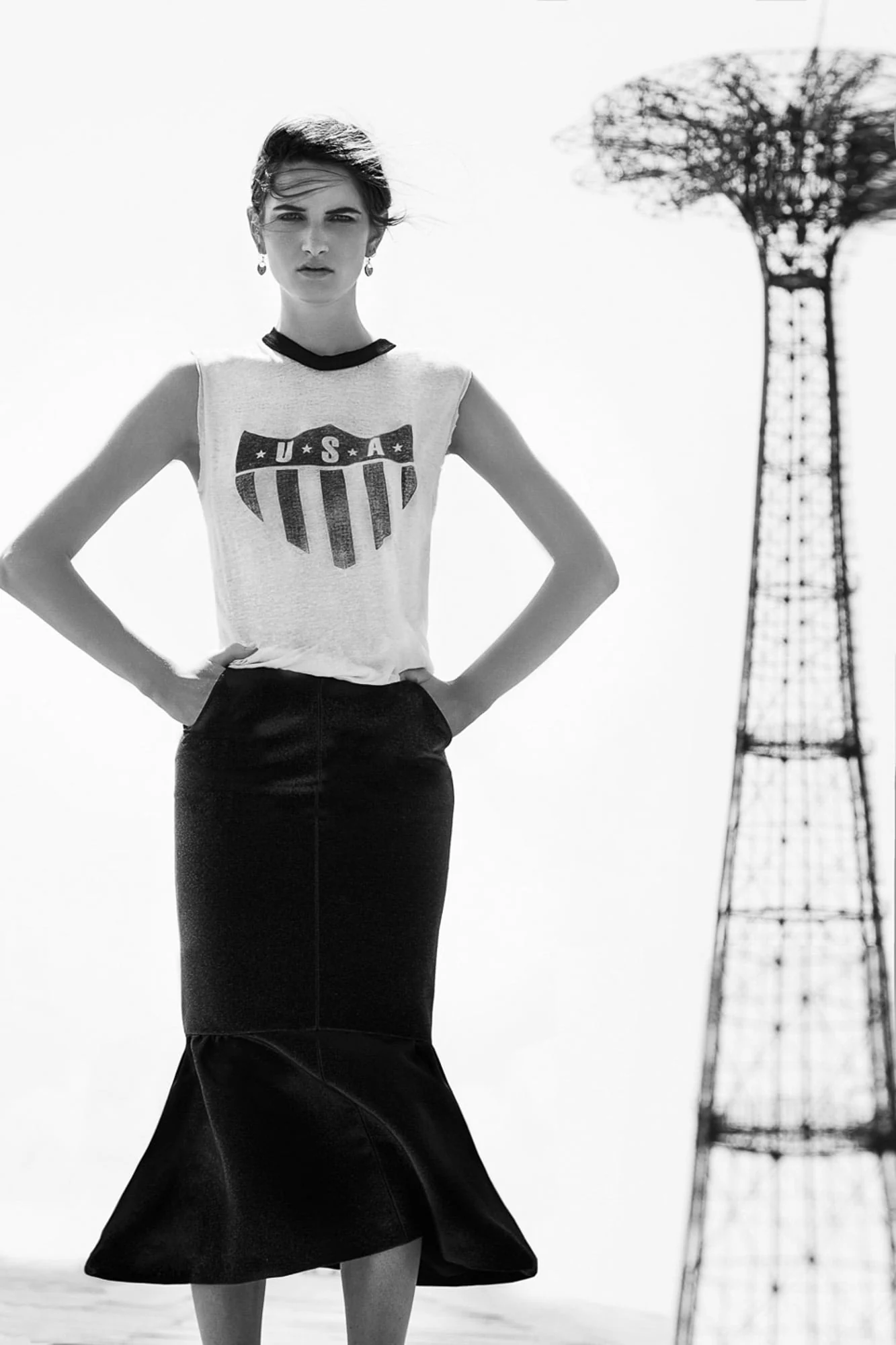 Black-and-white shot of a model in a USA shield tank and black flared skirt, hands on hips, beside the Coney Island Parachute Jump.