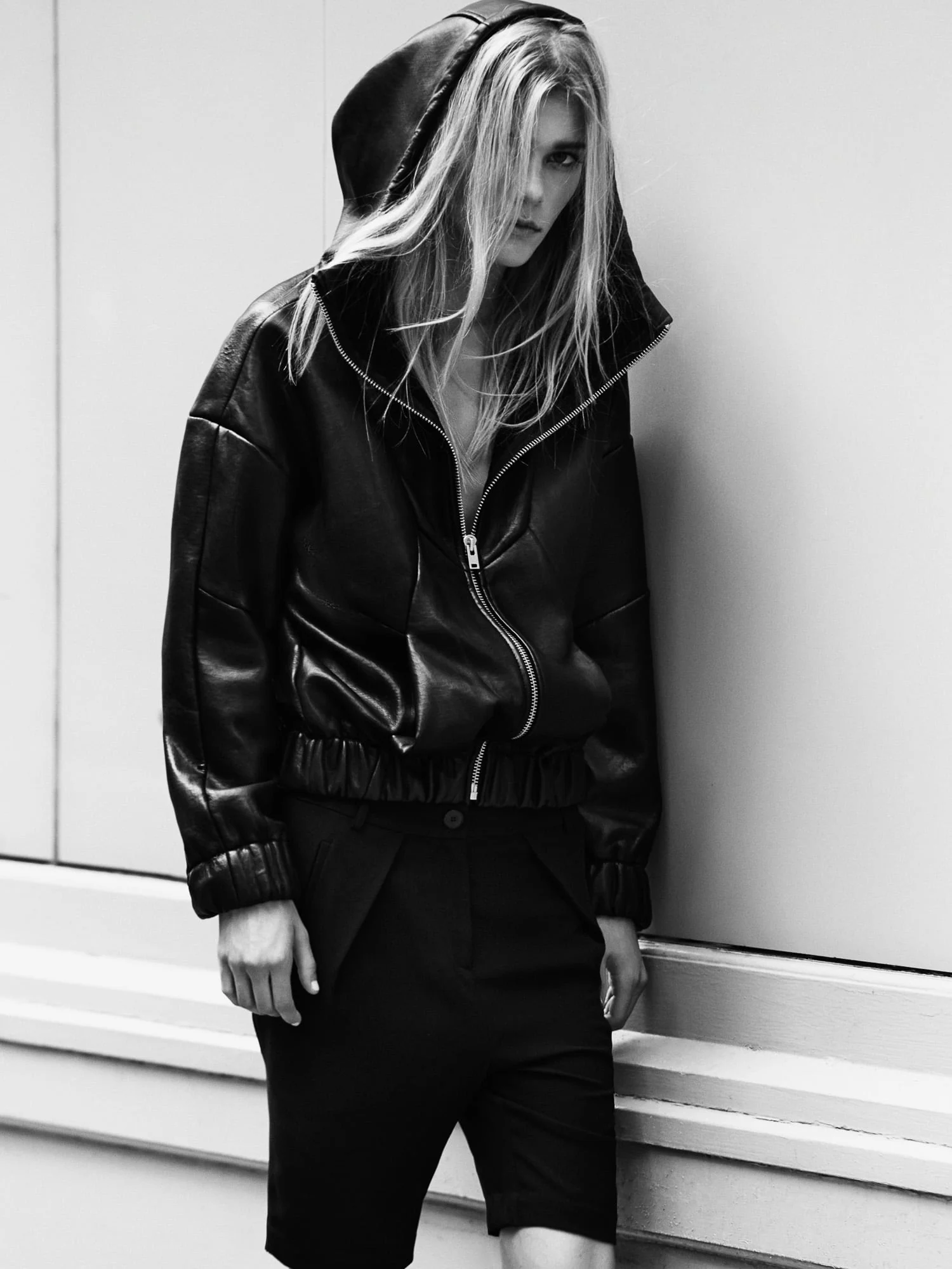 Black-and-white portrait of a blonde model in a hooded black leather bomber and black shorts leaning against a pale wall.
