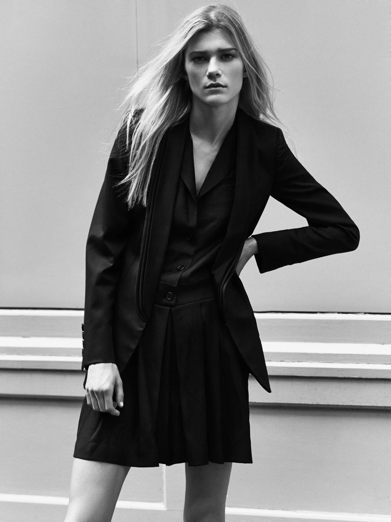 Black-and-white portrait of Cydnee Webb with long blonde hair in a black blazer and pleated skirt against a pale wall.