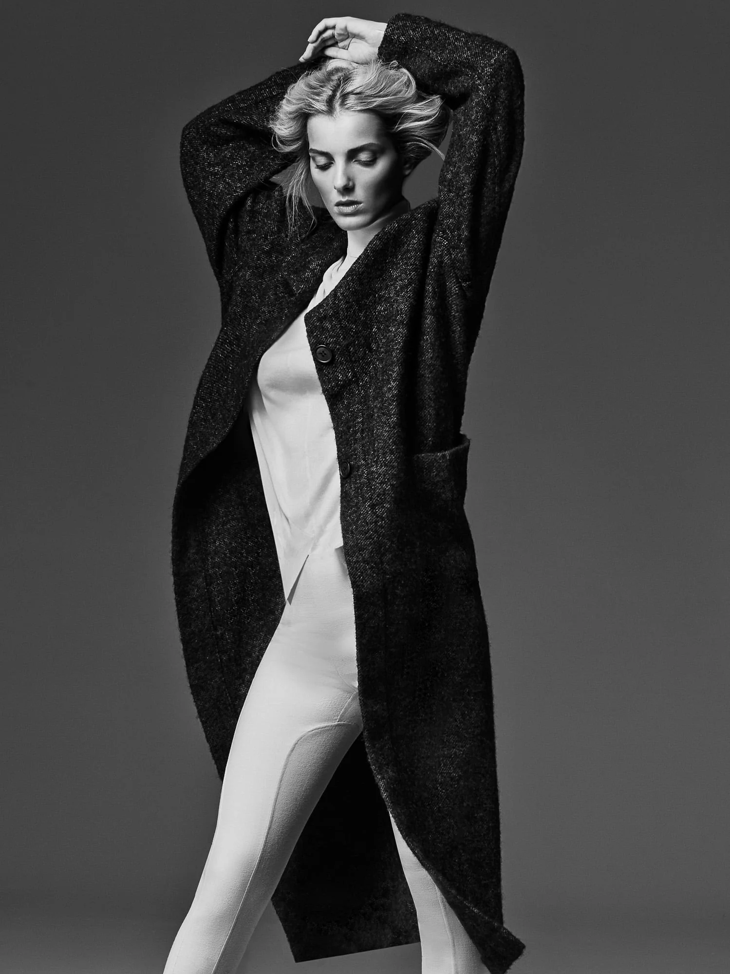 Black-and-white portrait of model Denisa Dvorakova in a long dark tweed coat over white, hands raised to her hair.