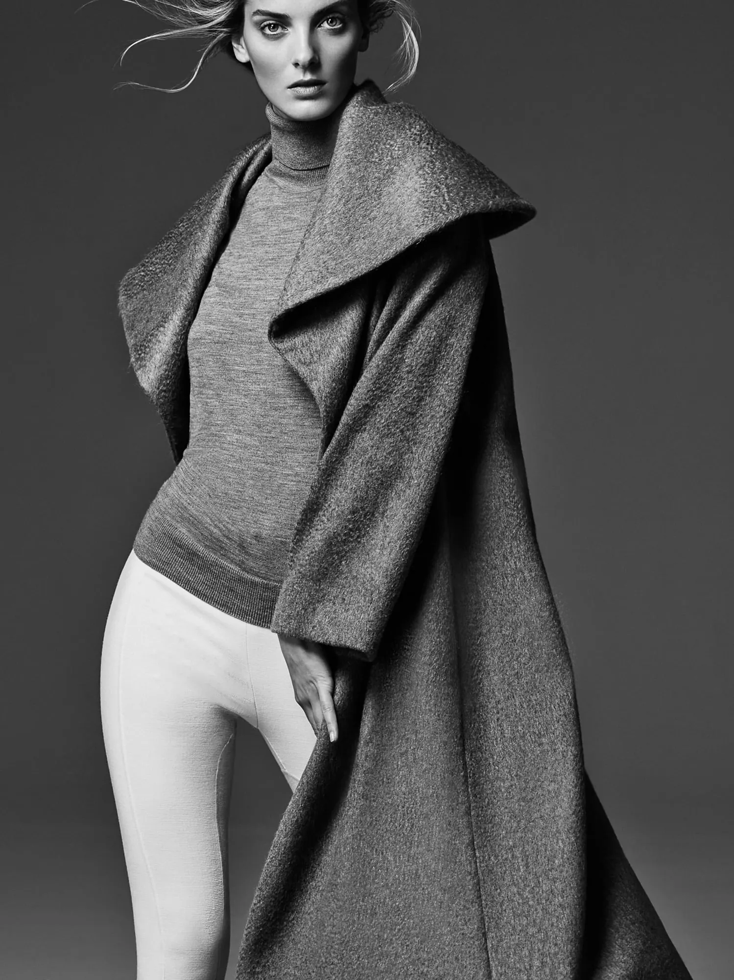 Black-and-white studio portrait of Denisa Dvorakova in a grey turtleneck, oversized wool coat and white trousers.