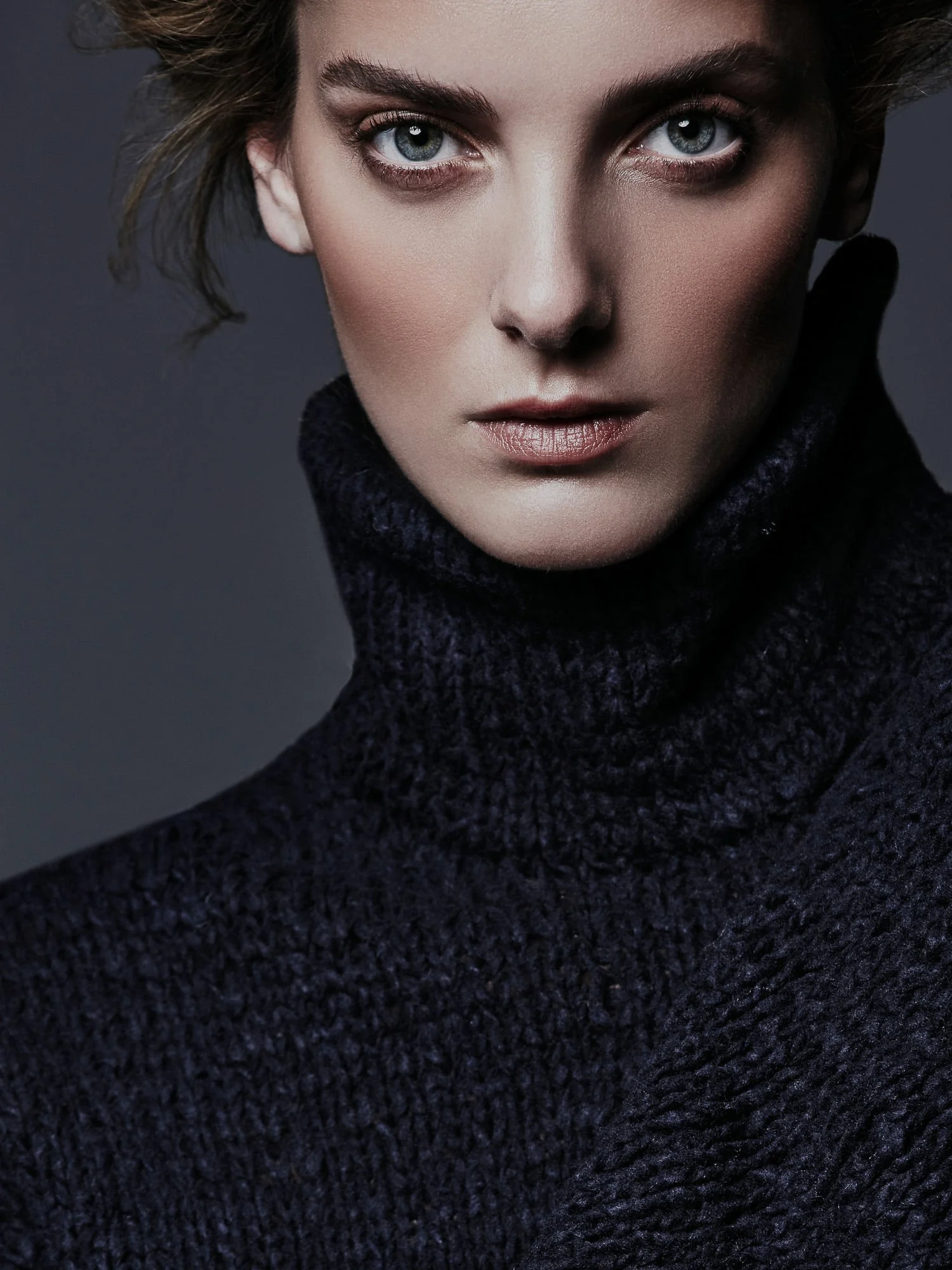 Close-up portrait of model Denisa Dvorakova in a chunky navy knit turtleneck against a dark grey backdrop.