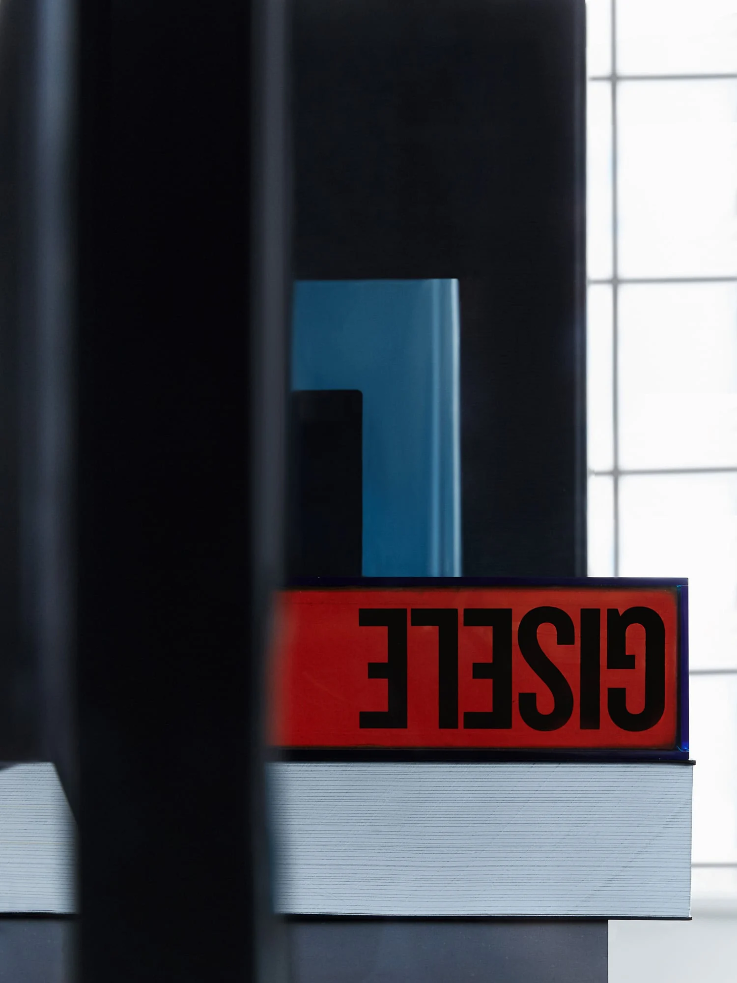 Stacked books with a red spine reading GISELE, a blue object and dark panels by a bright window, for Giovanni Bianco.