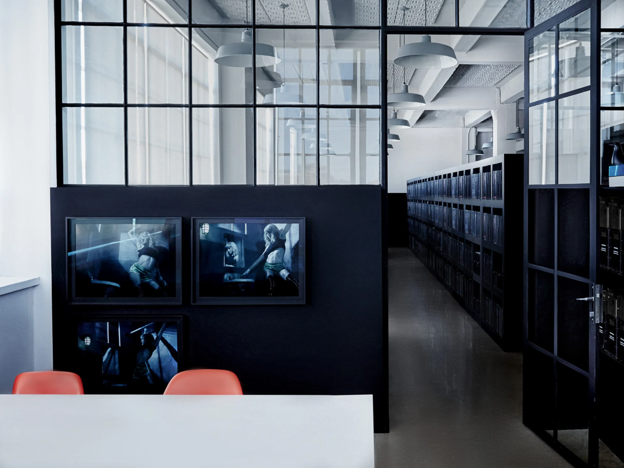 Office interior with black steel-framed glass partitions, framed photographs, orange chairs and archive shelving, for Giovanni Bianco.