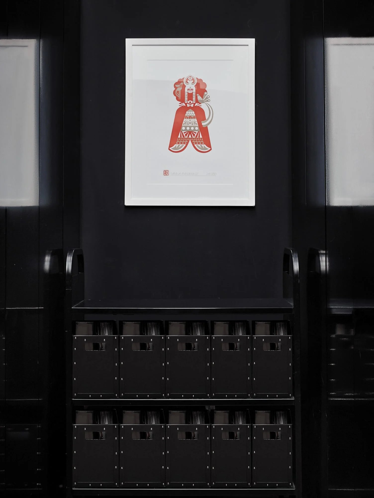Framed red folk-style figure print on a black wall above a black cart of storage boxes, for Giovanni Bianco.