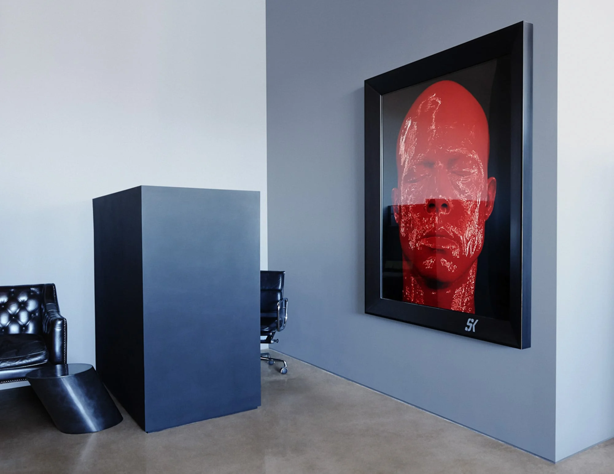 Office interior with a framed red glossy face artwork on a grey wall, black leather chair and dark pedestal, for Giovanni Bianco.
