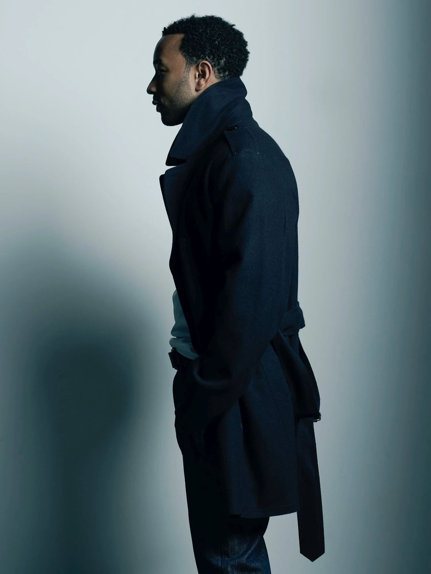 John Legend in profile wearing a navy belted wool coat against a pale grey studio wall with soft shadow.