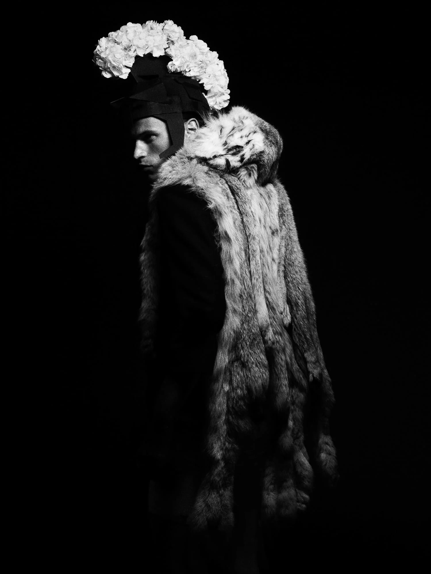 Black-and-white portrait of Lars Burmeister in a lynx fur coat and white floral headpiece, glancing over his shoulder.