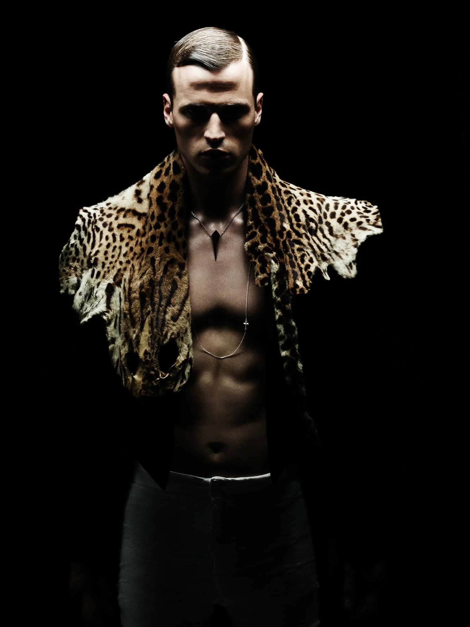 Lars Burmeister shirtless with a leopard-print pelt draped over his shoulders and a fang pendant, lit against black.