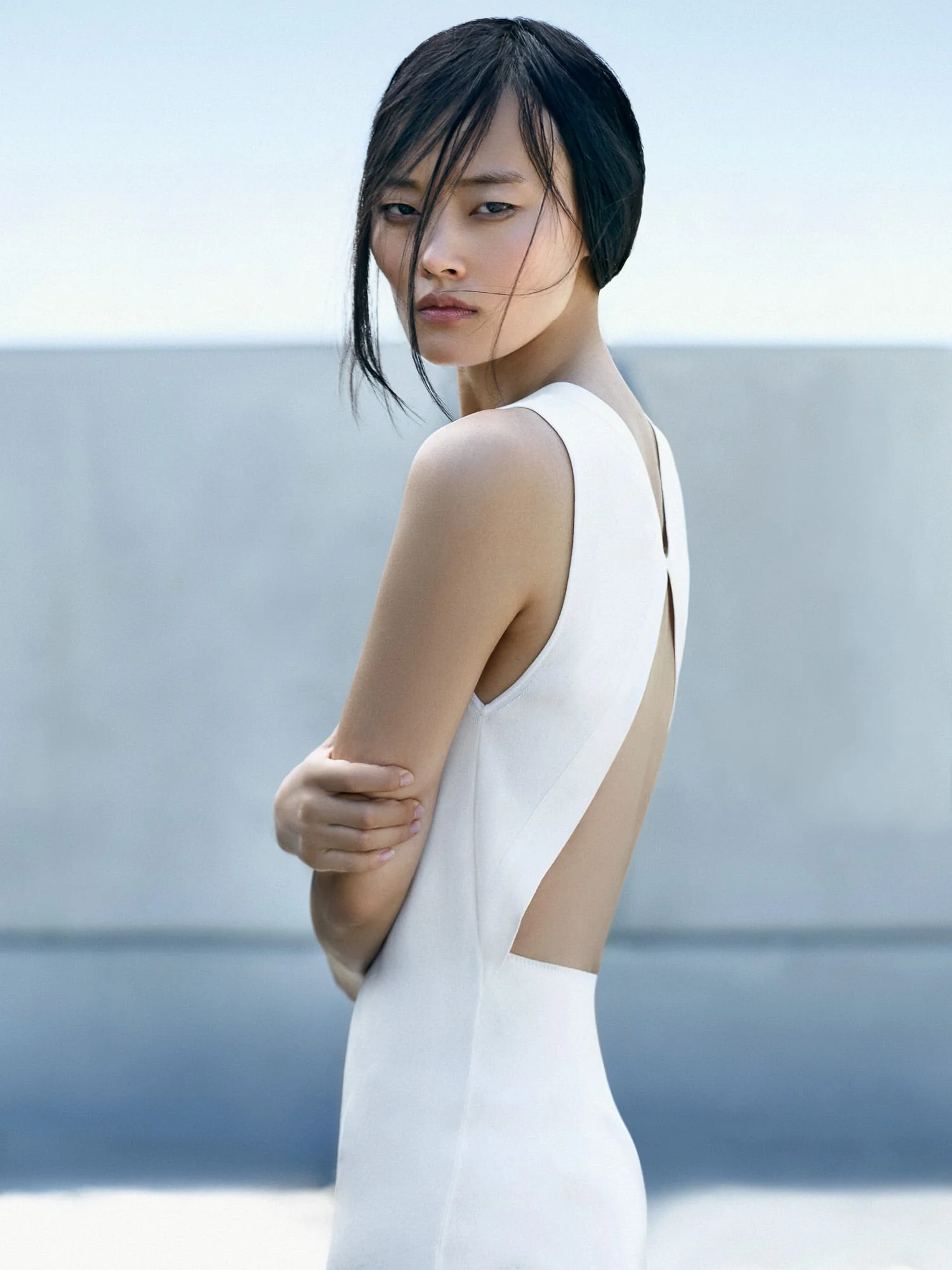 Model Li Xiang in a white open-back dress looking over her shoulder against a pale concrete wall and sky.