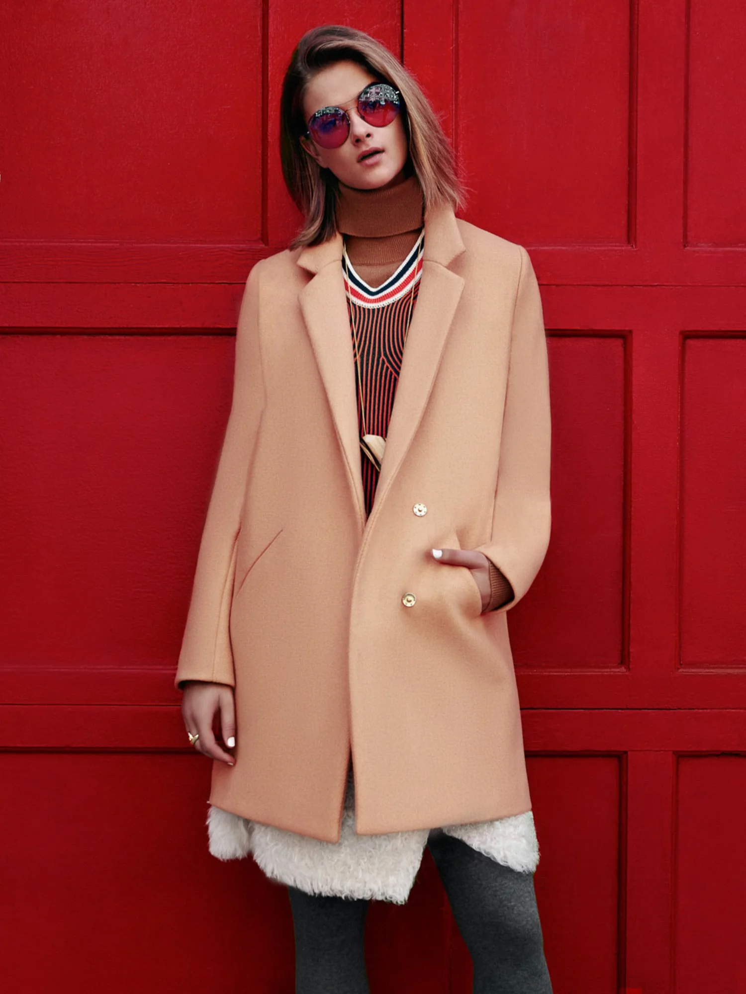 Model in a camel coat, striped knit and tinted round sunglasses posing against a red paneled door, for Lucky.