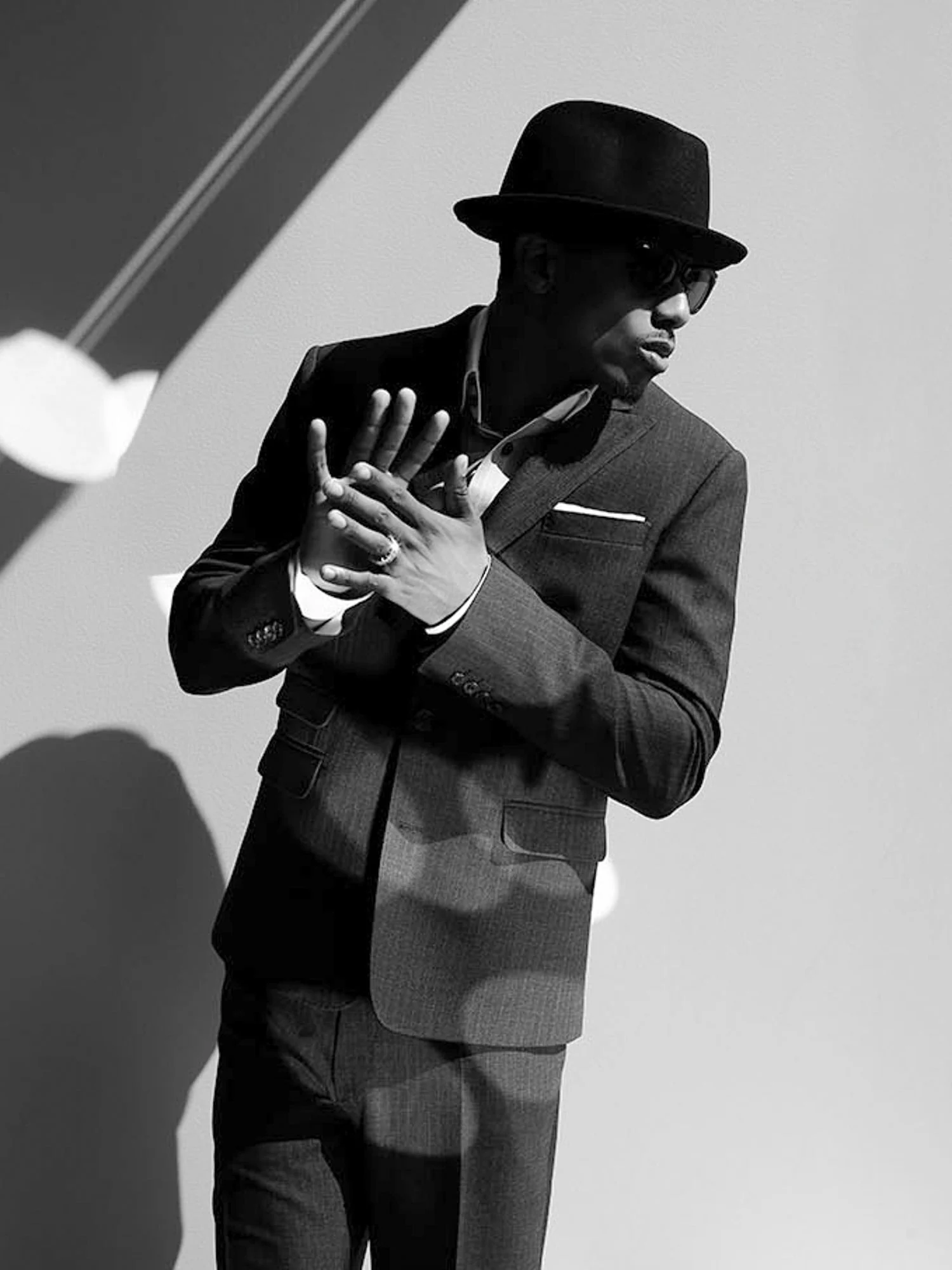Black-and-white portrait of Nick Cannon in a pinstripe suit, fedora and sunglasses, hands raised against a sunlit wall.