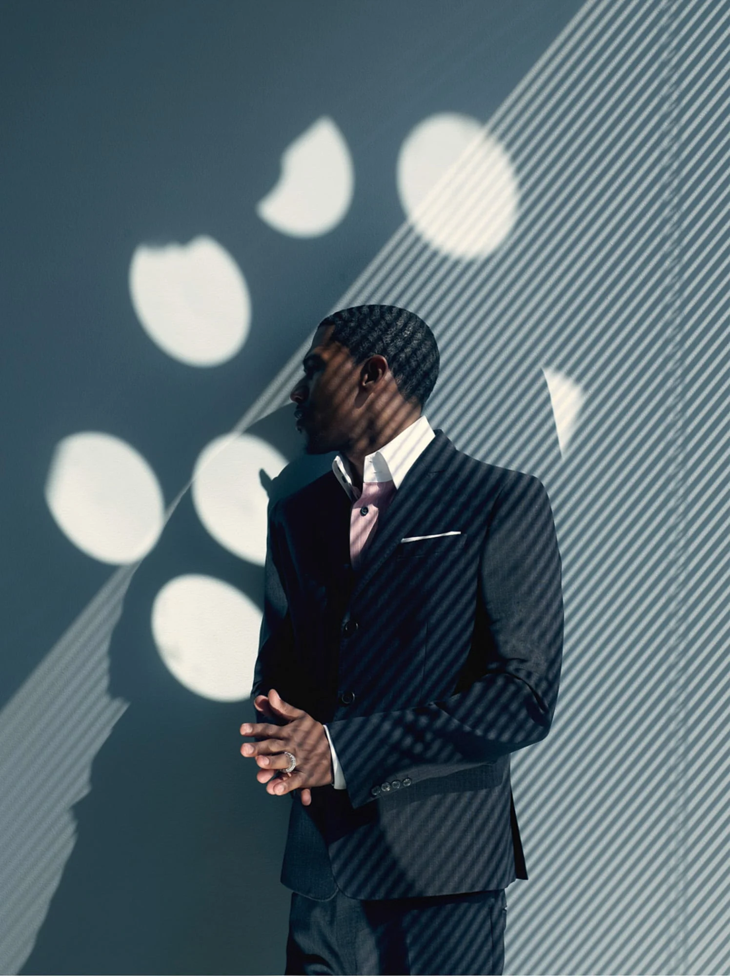 Nick Cannon in a navy suit and pink shirt, hands clasped, against a wall striped with blind shadows and circles of light. Photographed by Cameron Krone.
