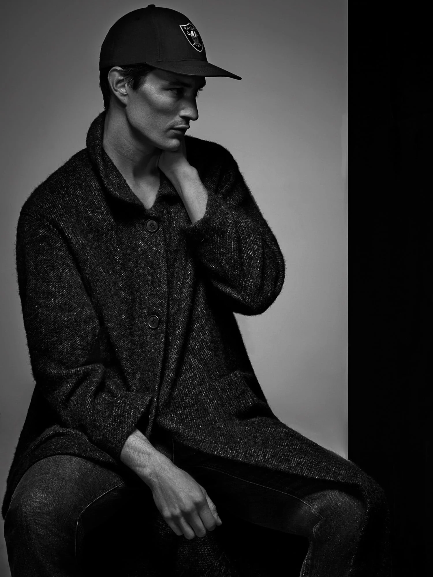 Black-and-white portrait of Owen Stuart seated in a dark tweed coat, jeans and baseball cap, hand on his neck.