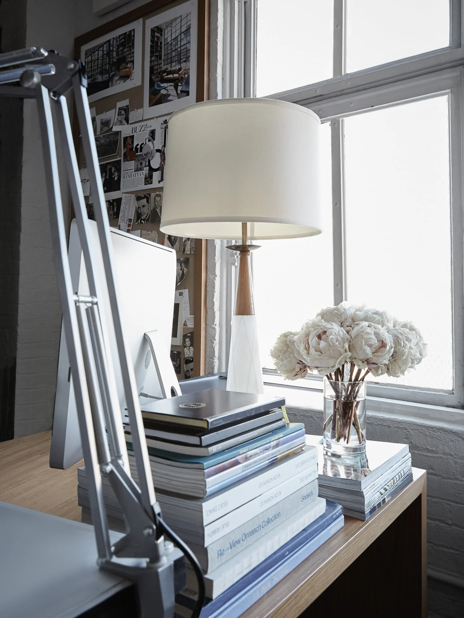 Desk by a loft window with a white table lamp, stacked books, a silver task lamp and a vase of white peonies, for Viana Art. Photographed by Cameron Krone.