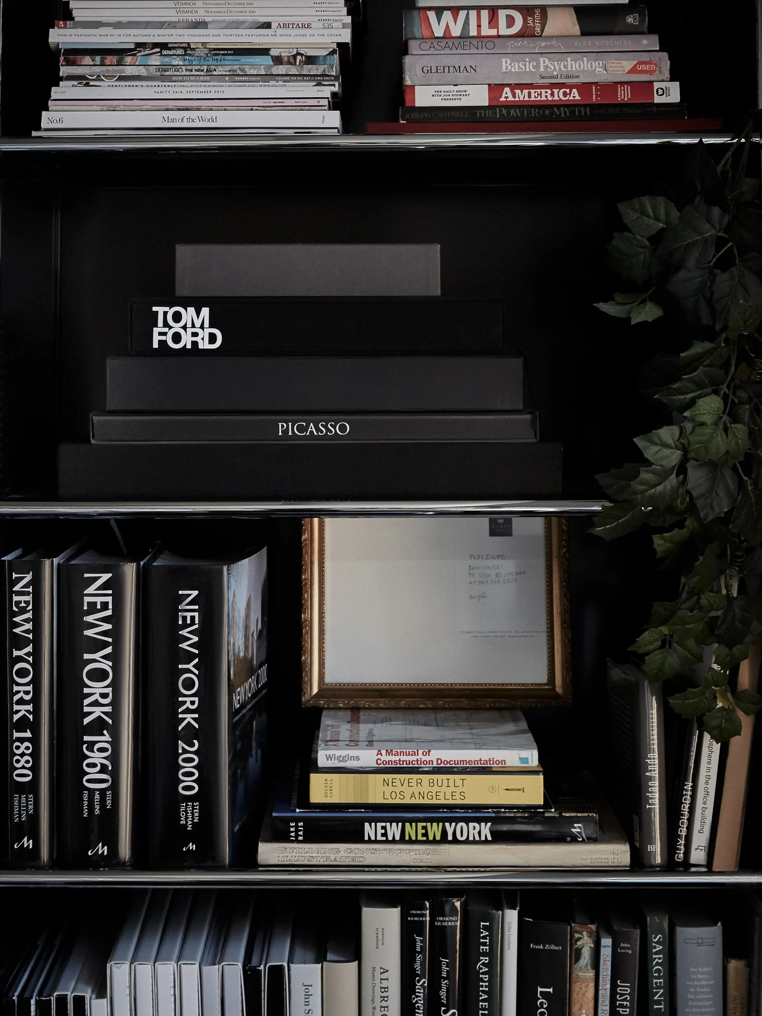 Black shelving with stacked art books, New York volumes, a framed letter and trailing ivy, for Viana Art.