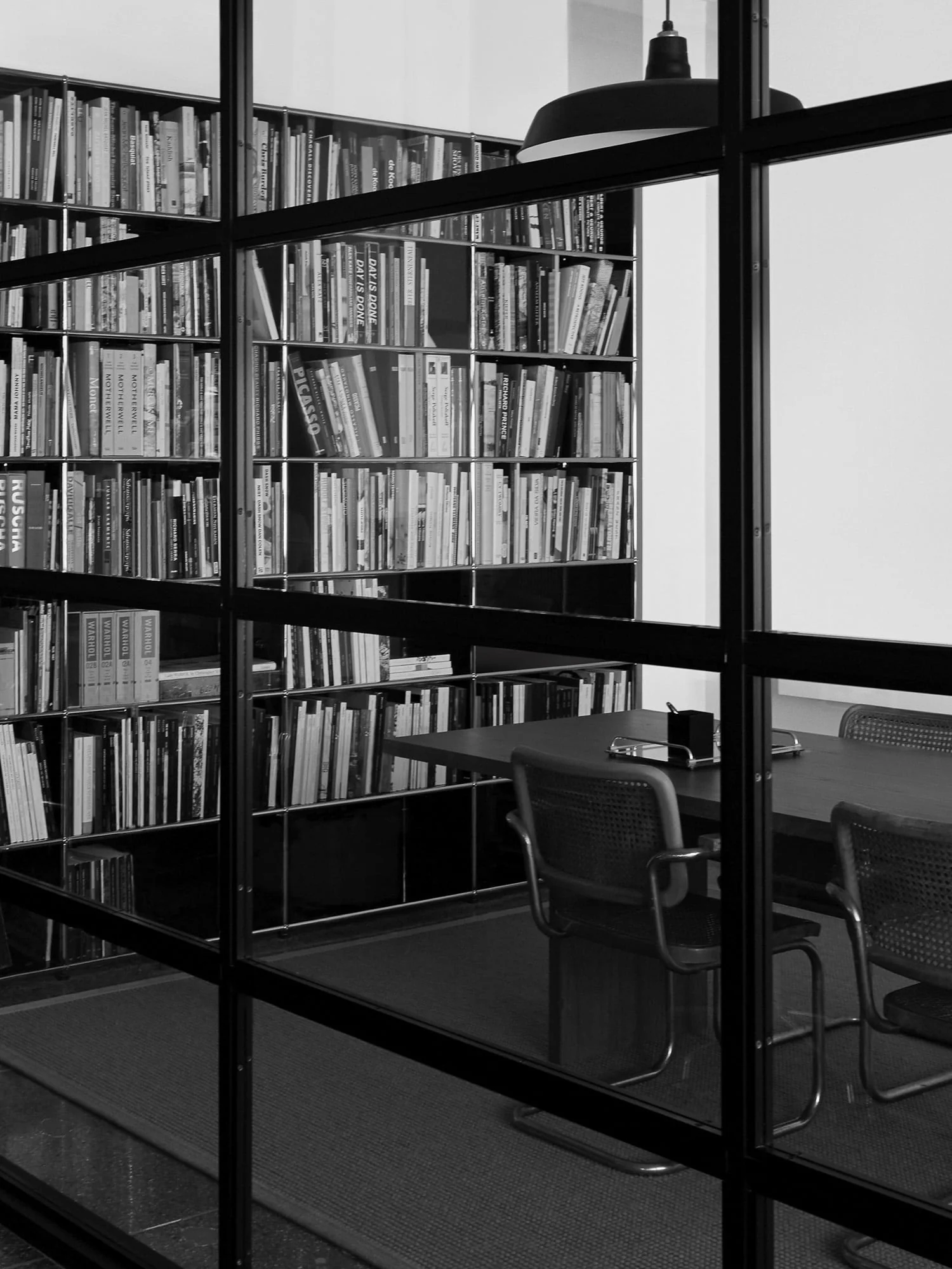 Black-and-white view through a steel-framed glass wall of tall bookshelves, a table and cane chairs, for Viana Art.