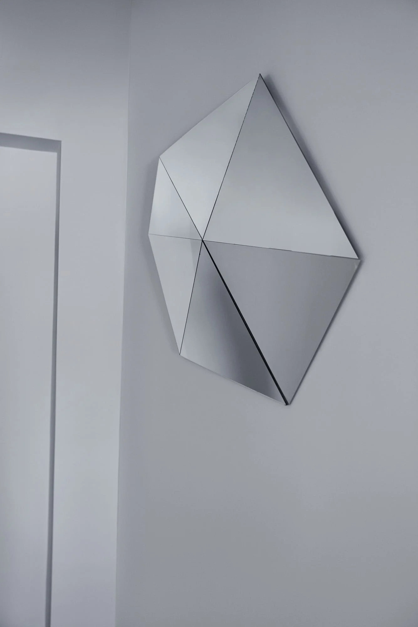 Faceted geometric mirror sculpture of triangular panels mounted on a pale grey wall beside a doorway, for Viana Art.