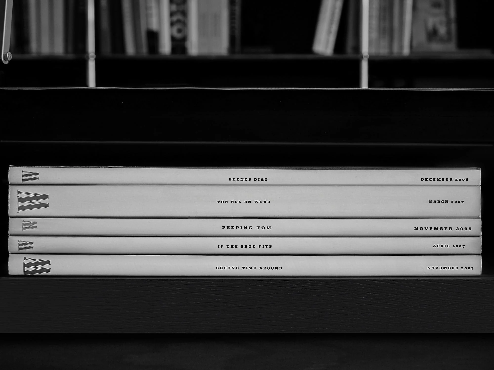 Black and white stack of five W magazine issues from 2005 to 2007, spines facing out on a dark bookshelf.