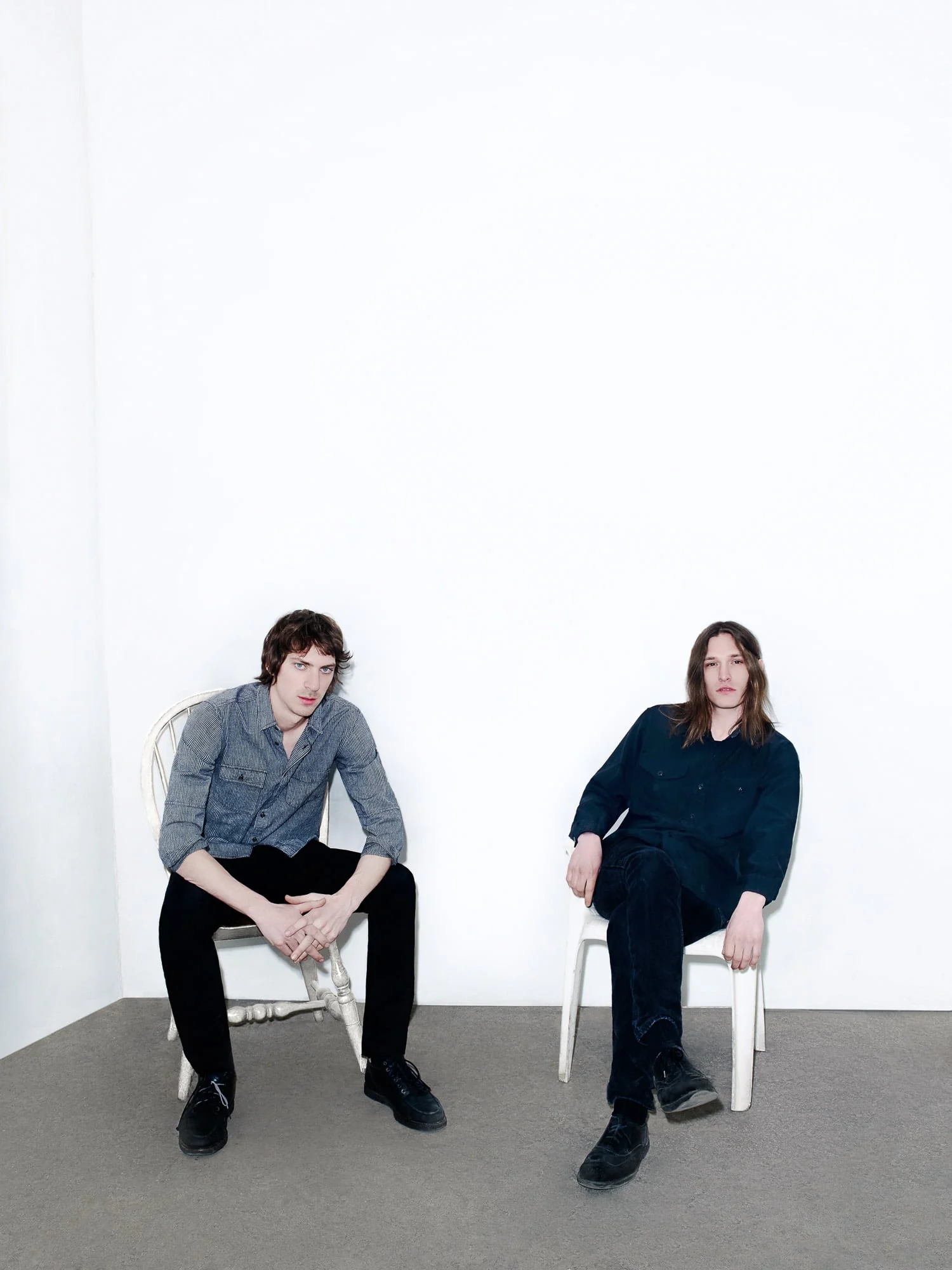 Two young men in dark shirts and black jeans seated on white chairs against a white studio wall, for The New York Times.