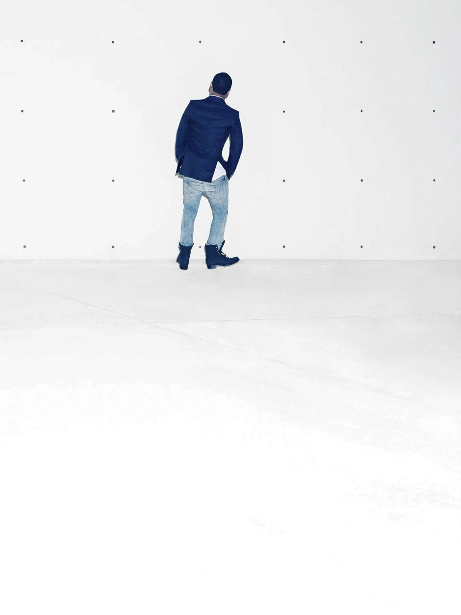 Man in navy blazer, pale jeans and dark boots stands facing a white concrete wall, for The New York Times.