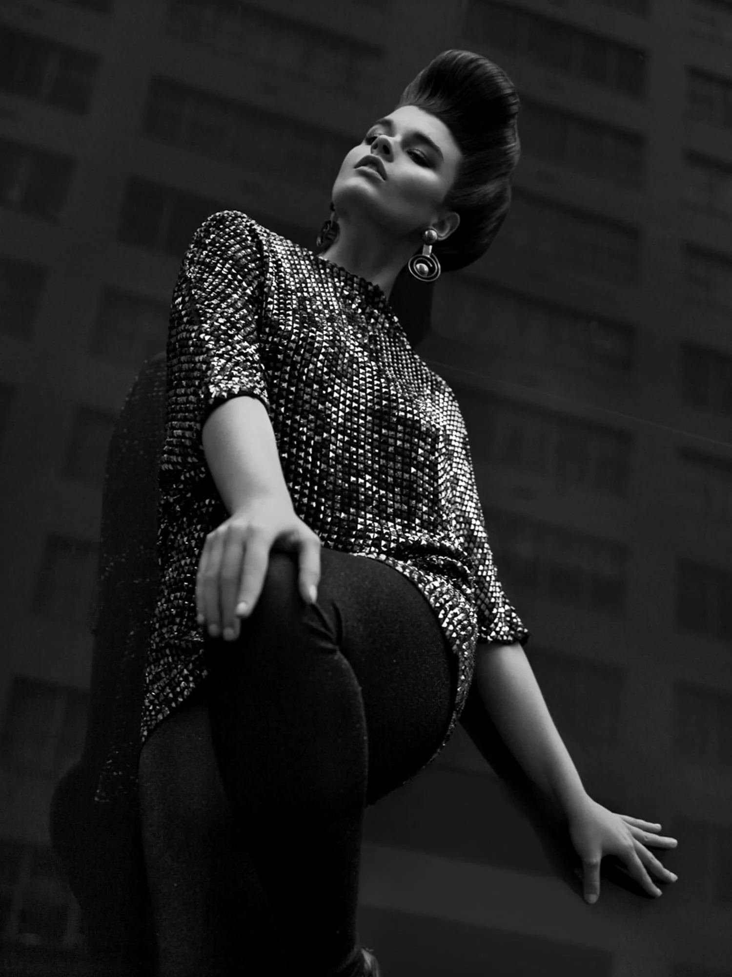 Black and white shot of a model with a high pompadour in a sequinned tunic and dark tights, posed against a tall building, for Sleek.