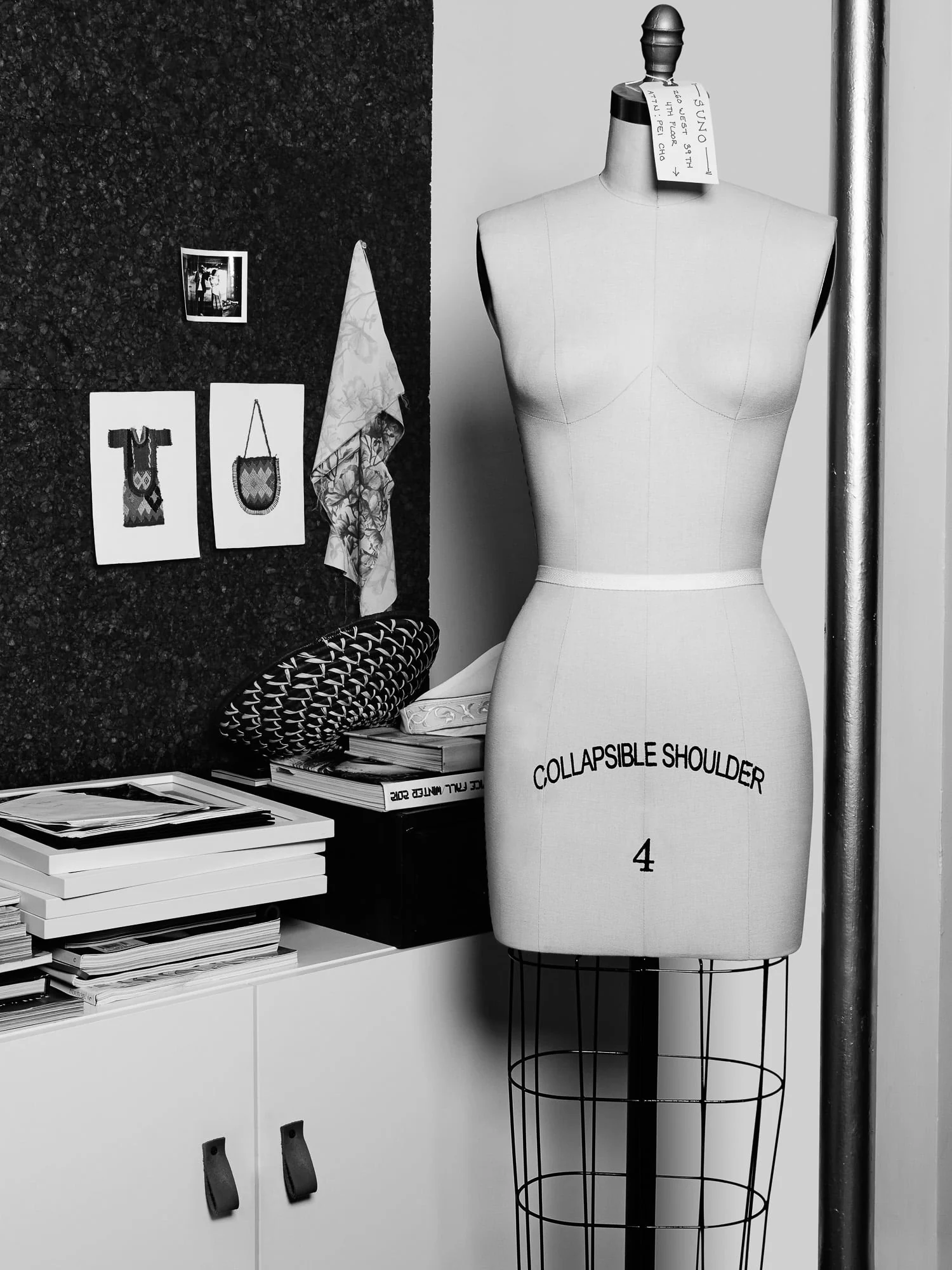 Black-and-white dress form tagged Suno beside a corkboard of sketches and stacked books in a design studio, for Elle.