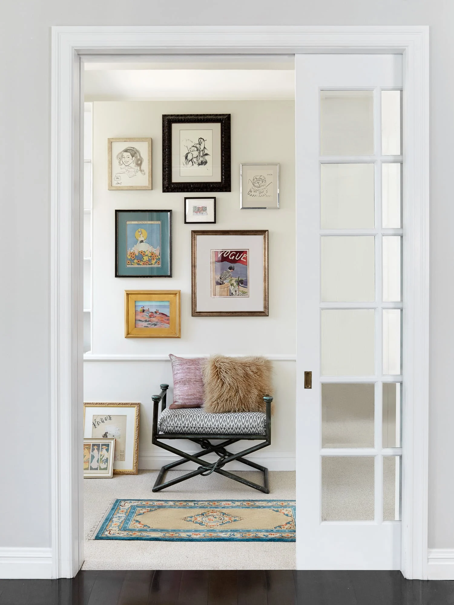 Gallery wall of framed vintage prints above a folding chair with furry cushions, seen through a white pocket door, for Harper's Bazaar.