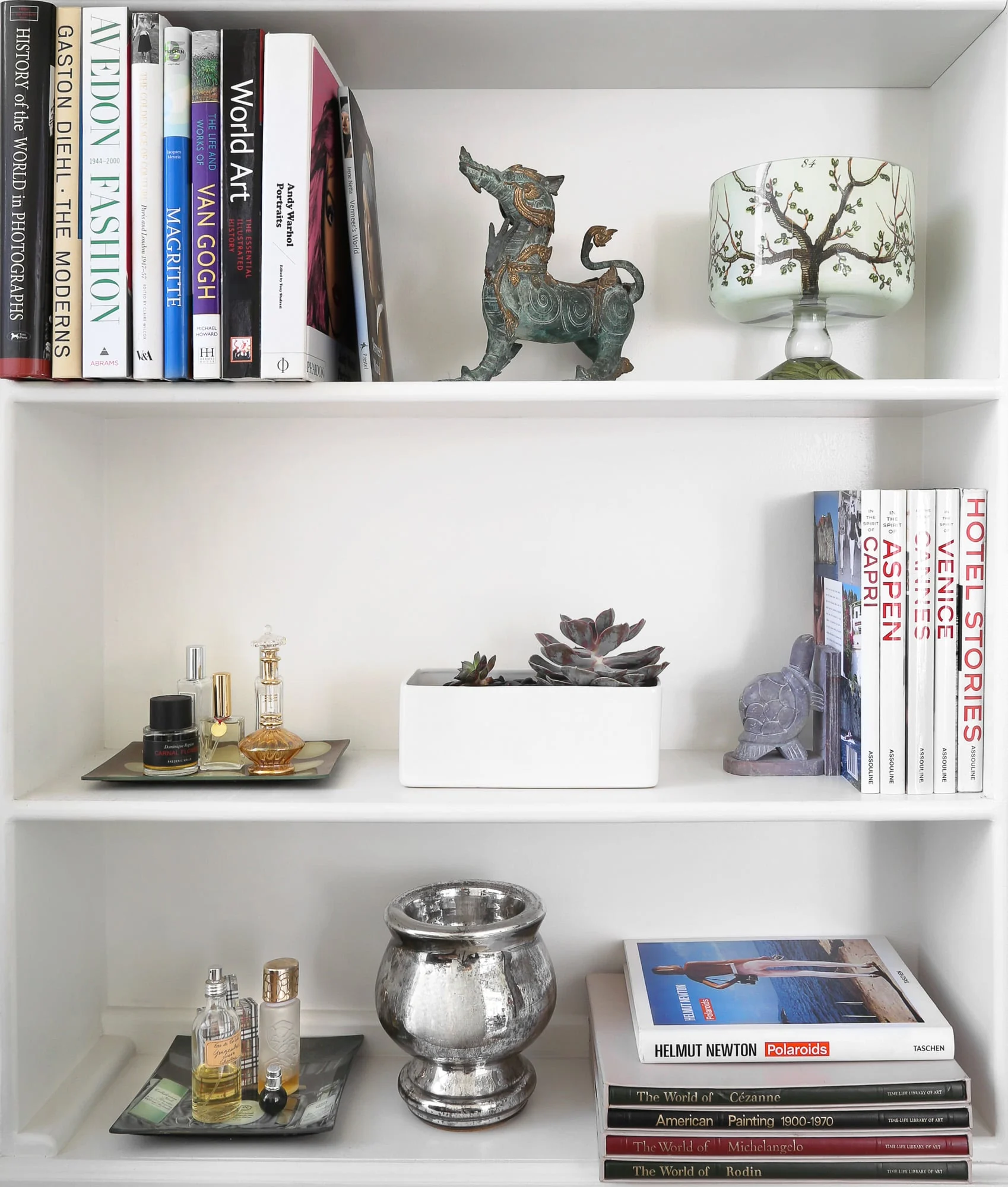 White bookshelf styled with art books, a bronze dragon figure, perfume bottles, a succulent and a silver vase, for Harper's Bazaar.