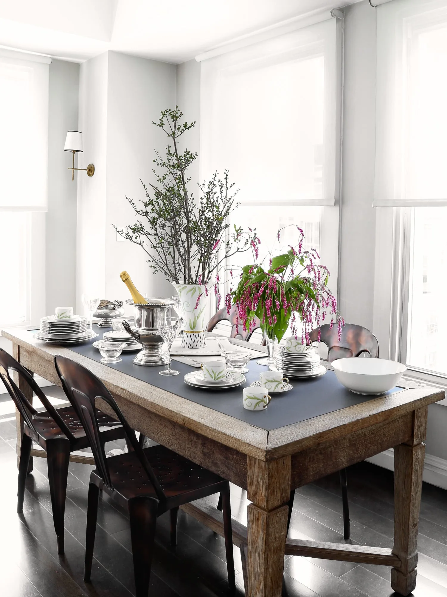Rustic wooden dining table set with white leaf-pattern porcelain, champagne bucket and pink flowers by bright windows, for Harper's Bazaar.