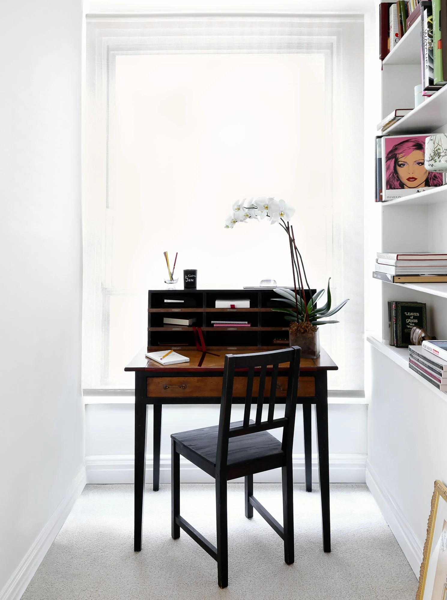 Small wooden writing desk with black chair, white orchid and letter organizer beside white bookshelves, for Harper's Bazaar.