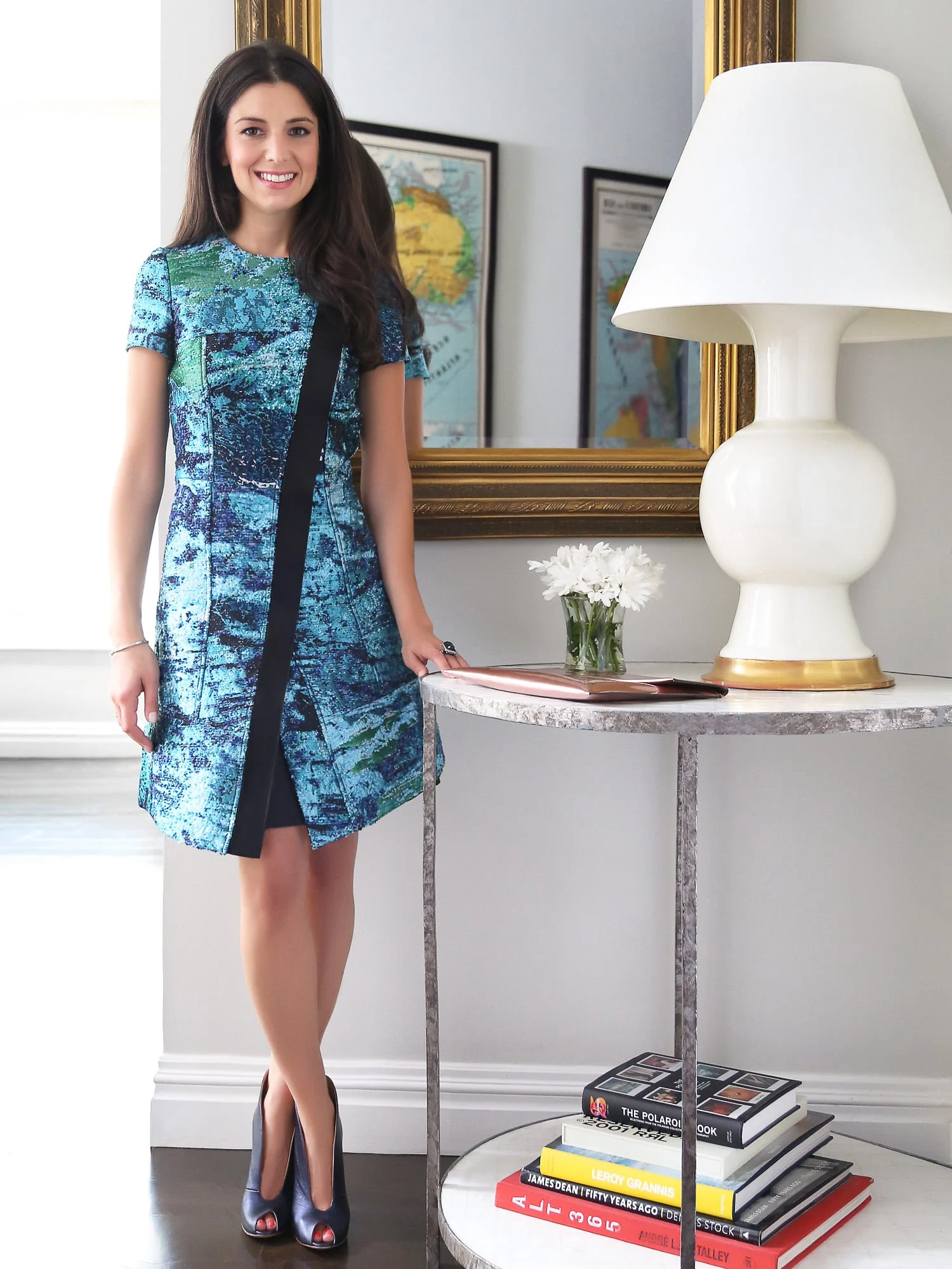 Smiling woman in a blue-green jacquard dress beside a marble side table, white lamp and gilt mirror, for Harper's Bazaar.