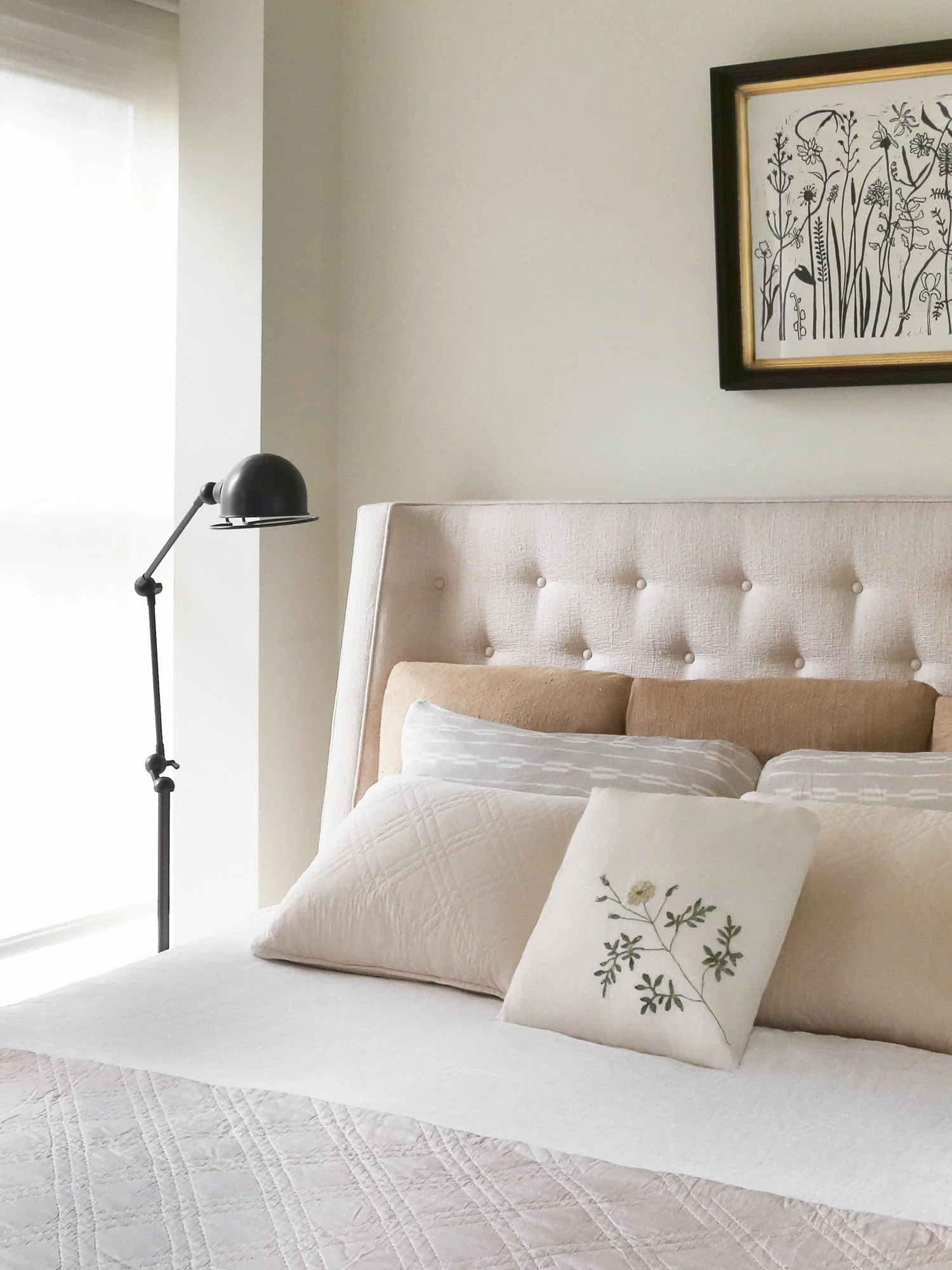 Bed with tufted linen headboard, cream pillows, embroidered cushion, black floor lamp and botanical print, for Harper's Bazaar.