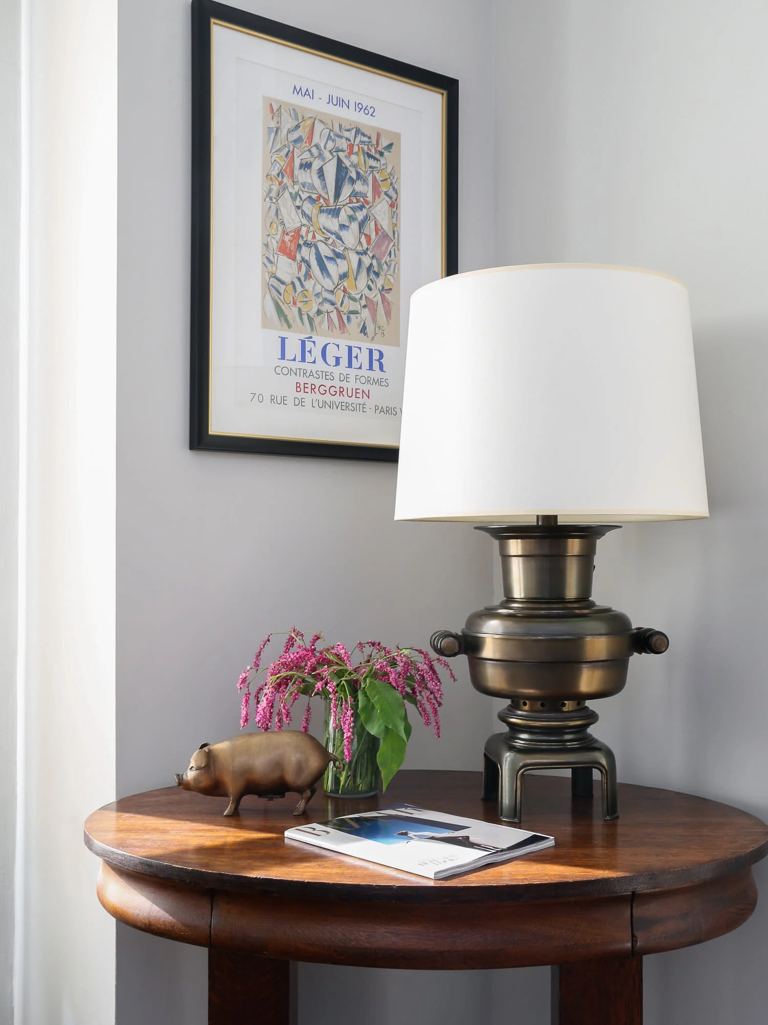 Round wooden side table with brass lamp, pink flowers, brass pig and magazine under a framed Léger poster, for Harper's Bazaar. Photographed by Cameron Krone.