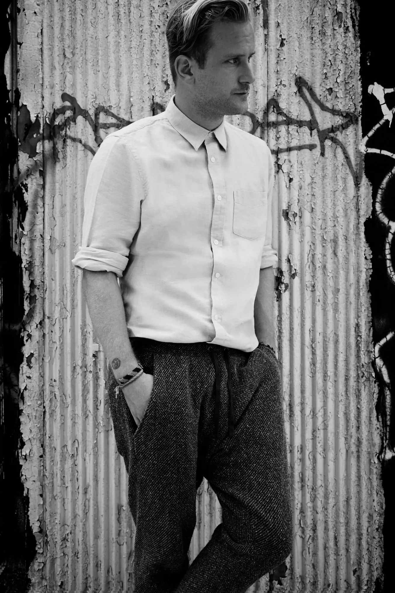 Black-and-white shot of a man in a white shirt and tweed trousers leaning on a graffitied corrugated wall, for Hercules.