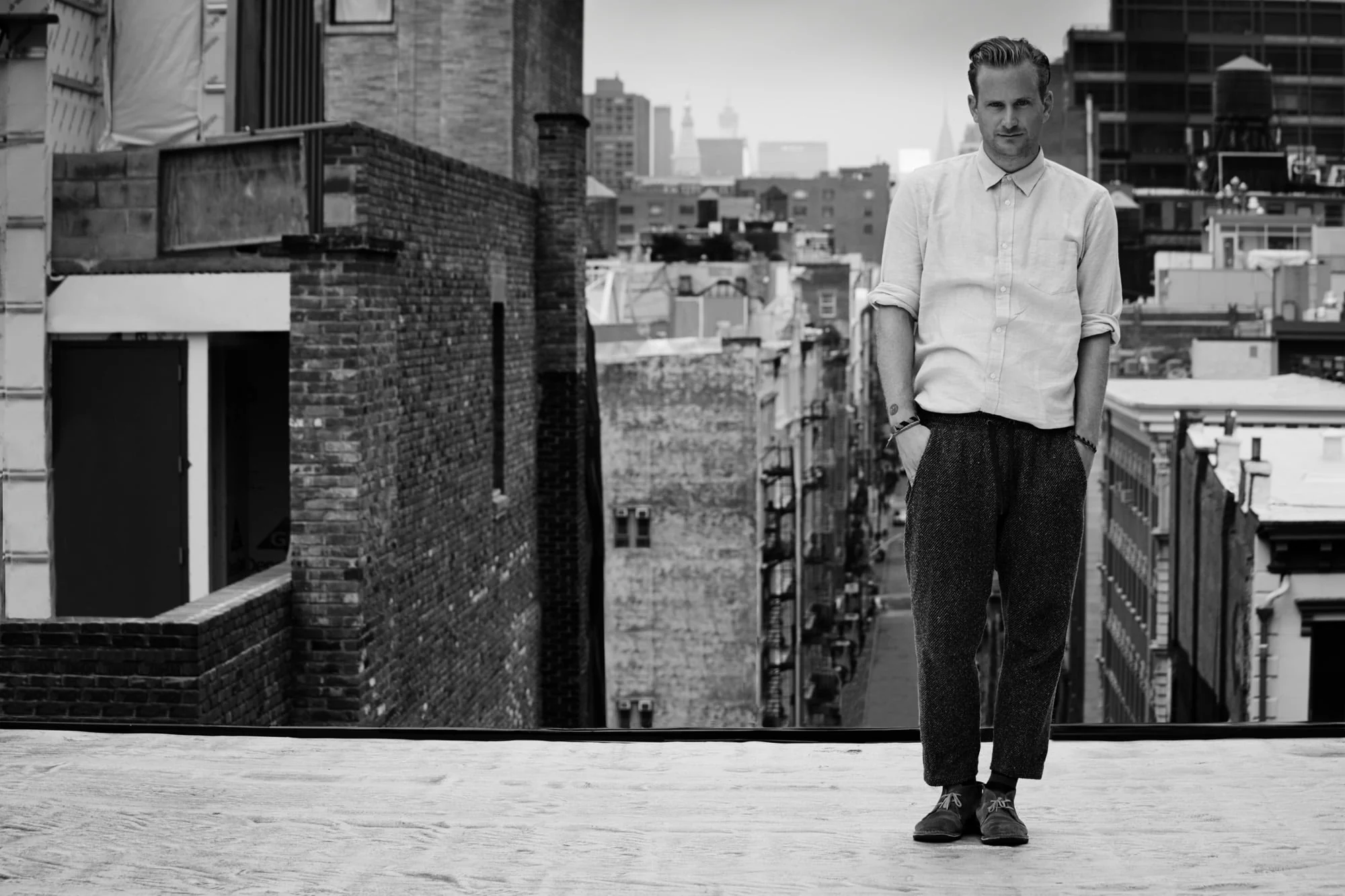Black-and-white portrait of designer Robert Geller standing on a Manhattan rooftop in a pale shirt and wool trousers.