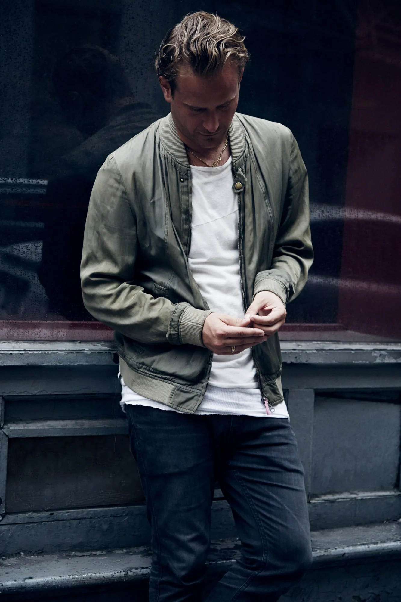 Man in an olive bomber jacket, white tee and dark jeans leaning against a storefront, wearing Robert Geller, for Hercules.