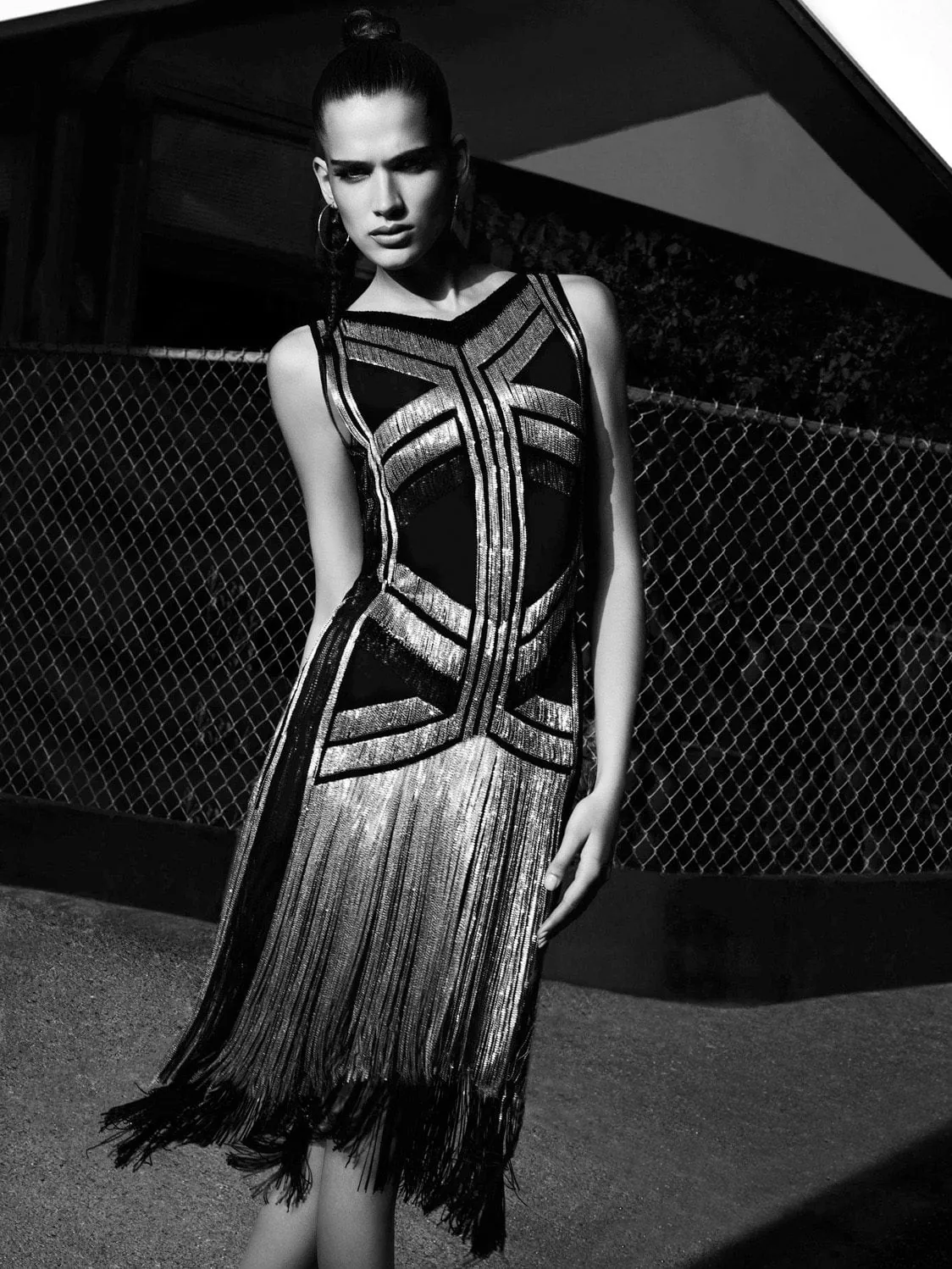 Black-and-white shot of a model in a black dress with metallic bands and fringe by a chain-link fence, for Amica.