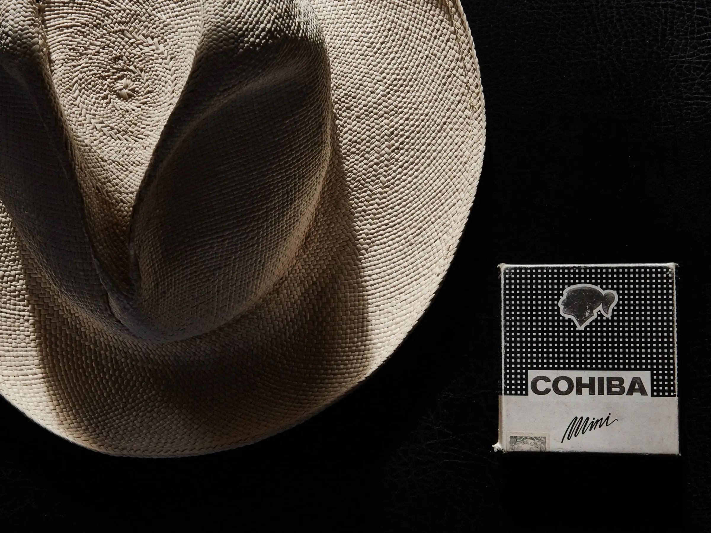 Woven straw Panama hat beside a box of Cohiba Mini cigars on black leather, still life, for Jeff Rudes.