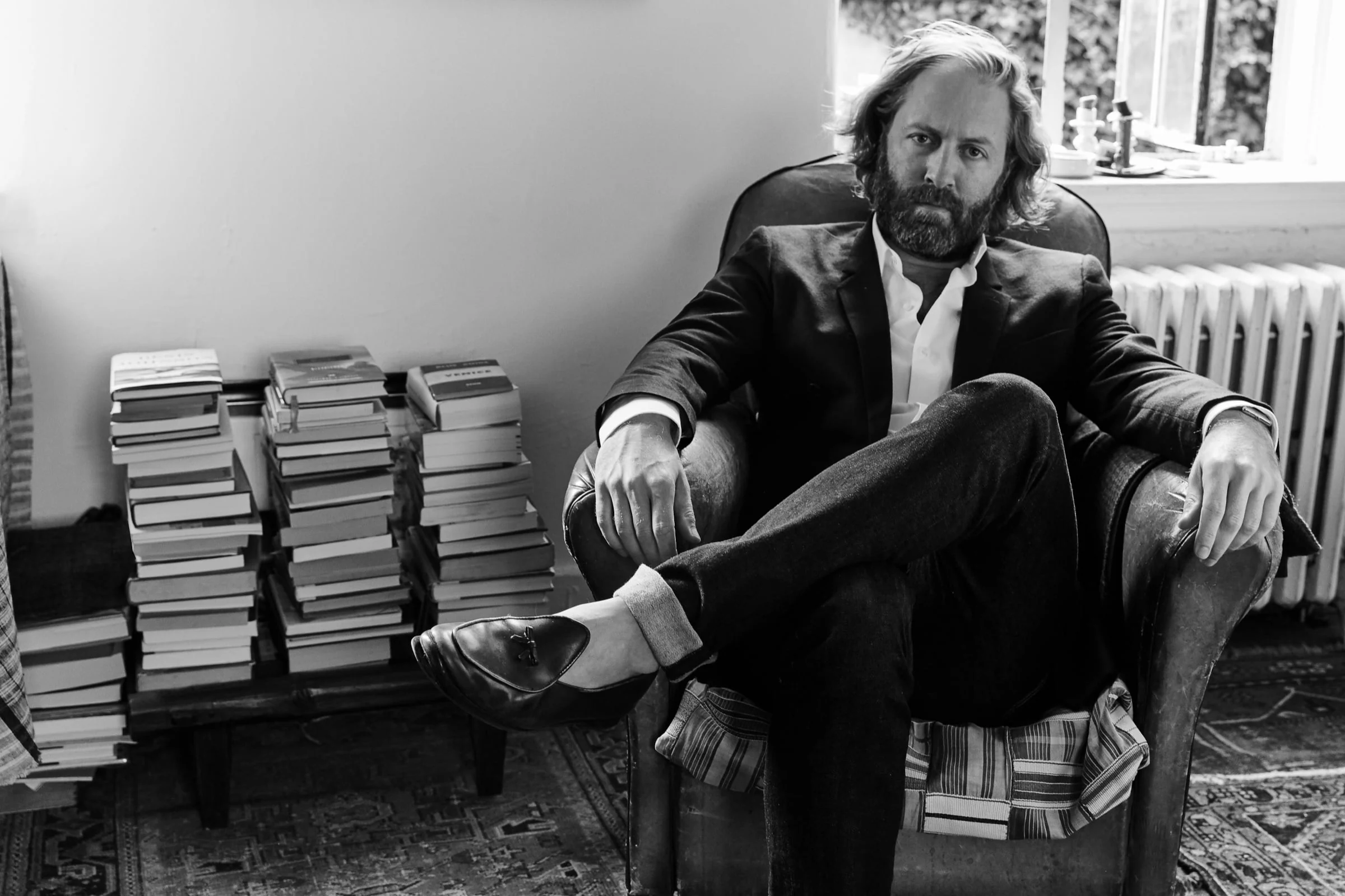 Black and white portrait of Jeff Rudes seated in a worn leather armchair beside stacks of books and a window.