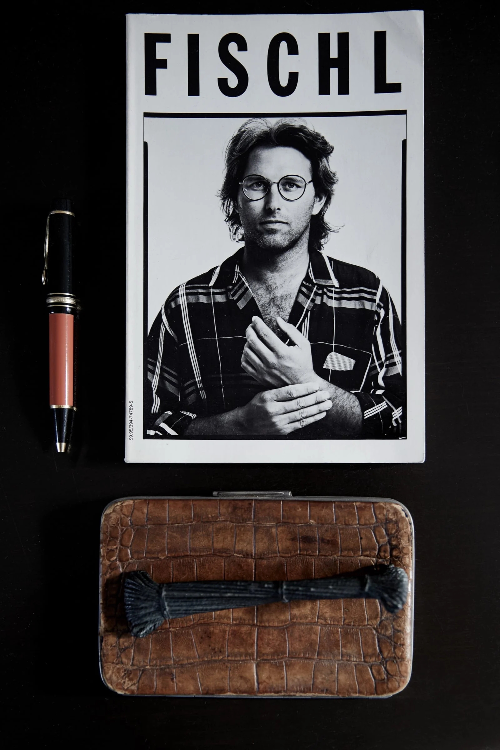 Still life of a Fischl book with a black and white portrait cover, a fountain pen and a crocodile leather box on dark wood.