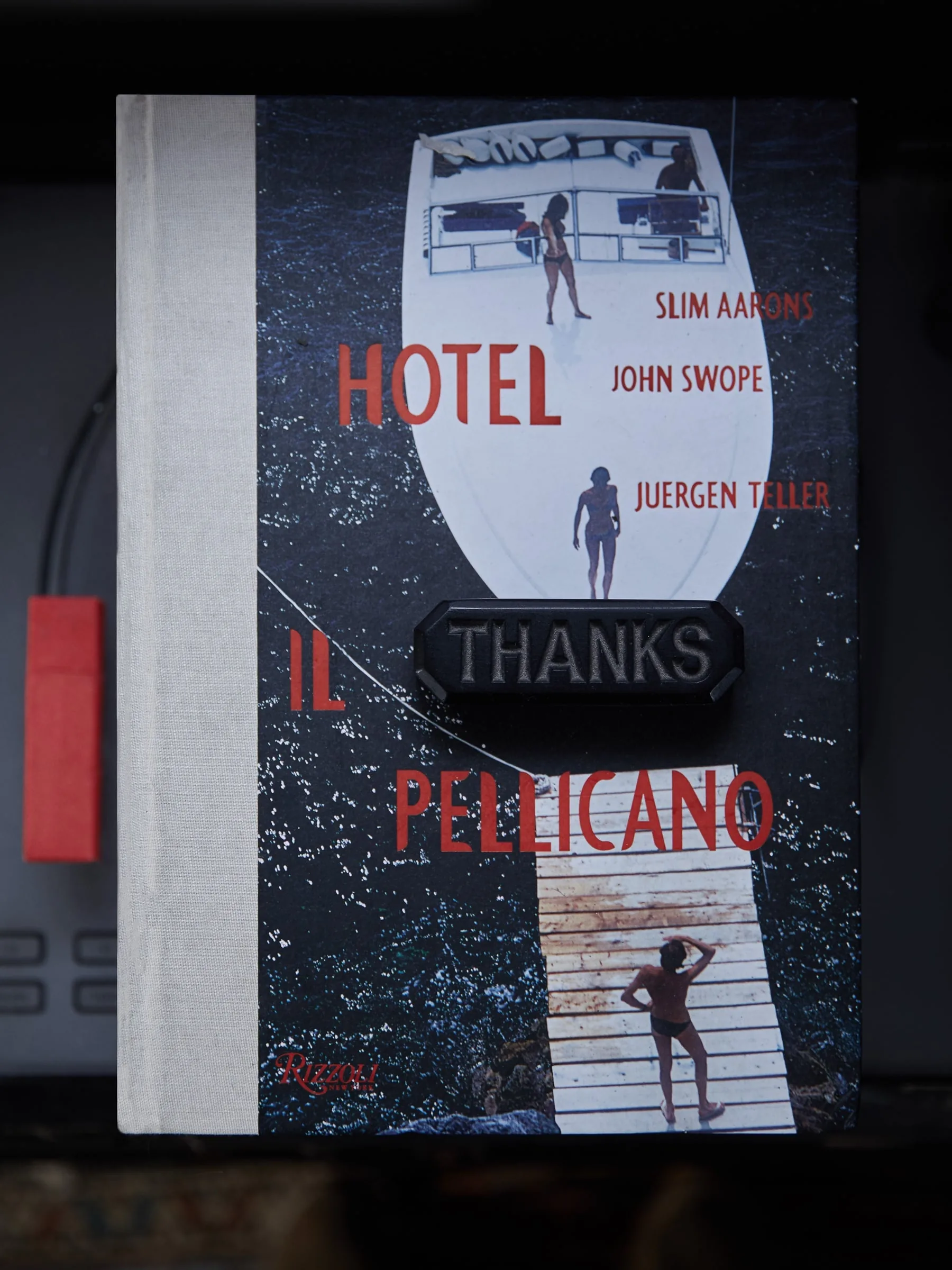 Hotel Il Pellicano book from Rizzoli with a black "Thanks" plaque on its cover, resting on a dark shelf.